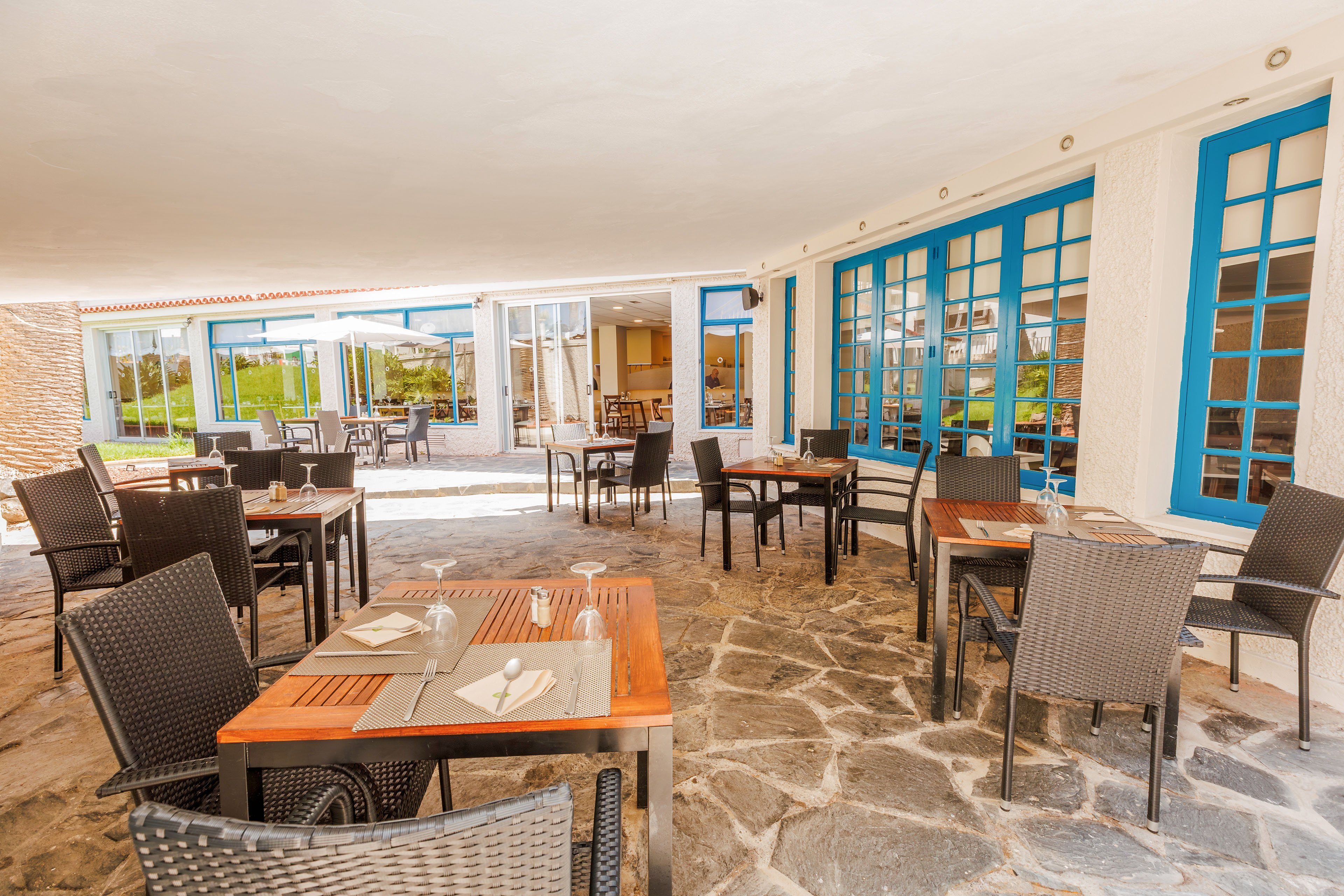 An open-air restaurant terrace features several wooden tables with wicker chairs arranged on a stone floor. Neutral-colored placemats and neatly folded napkins are set on the tables, ready for guests. Large blue-framed windows and doors line the building, adding a bright accent to the warm, sunlit space. In the background, the interior dining area and a glimpse of greenery are visible through the glass doors.