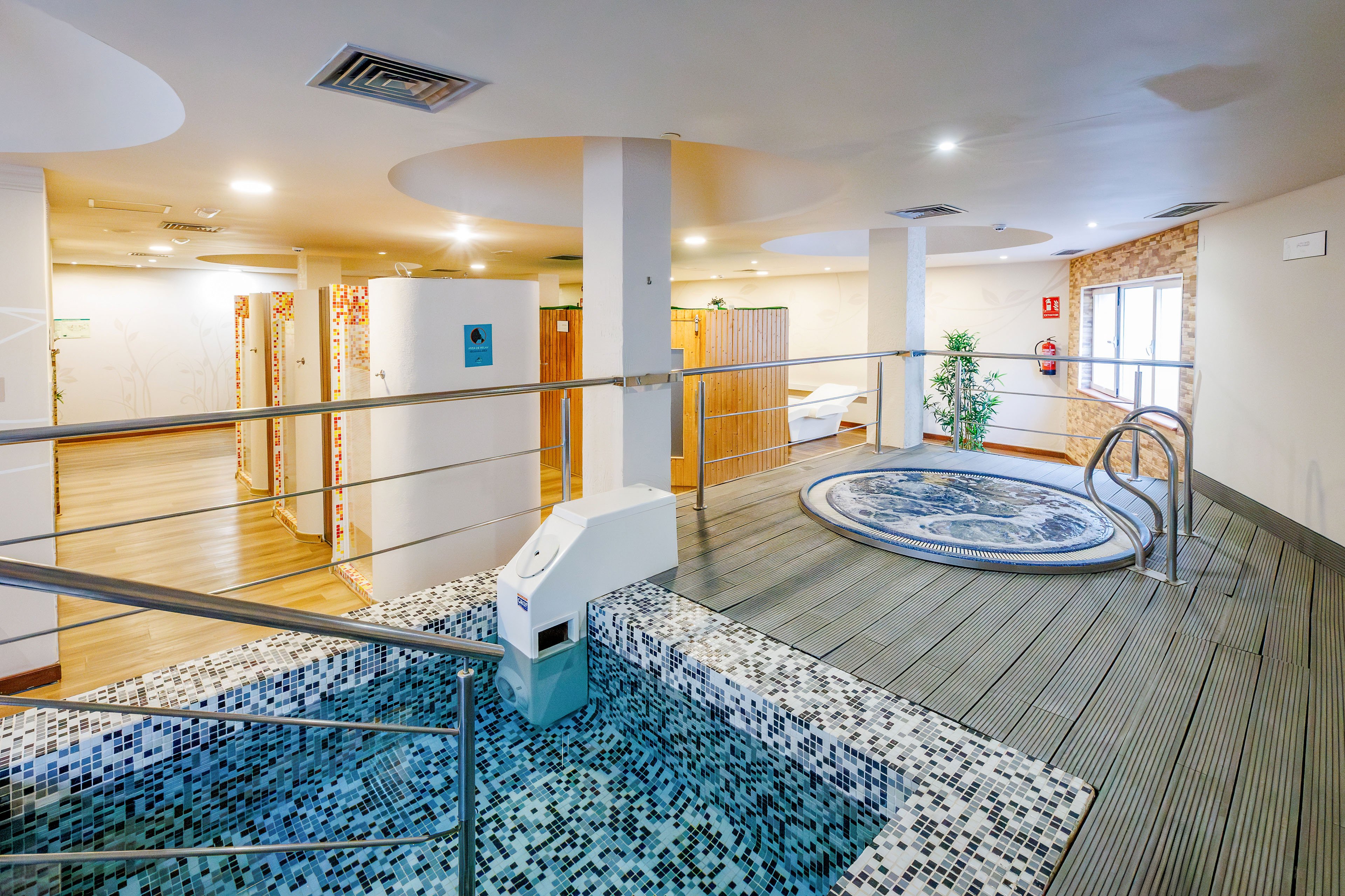 A contemporary indoor spa area features a small tiled pool in the foreground and a circular jacuzzi in the background. The space is finished with light wood flooring, white walls, and recessed ceiling lights, creating a clean and relaxing atmosphere. Stainless steel railings and steps lead into the water, while a few plants and seating areas add comfort. The overall design emphasizes wellness, relaxation, and modern leisure facilities.