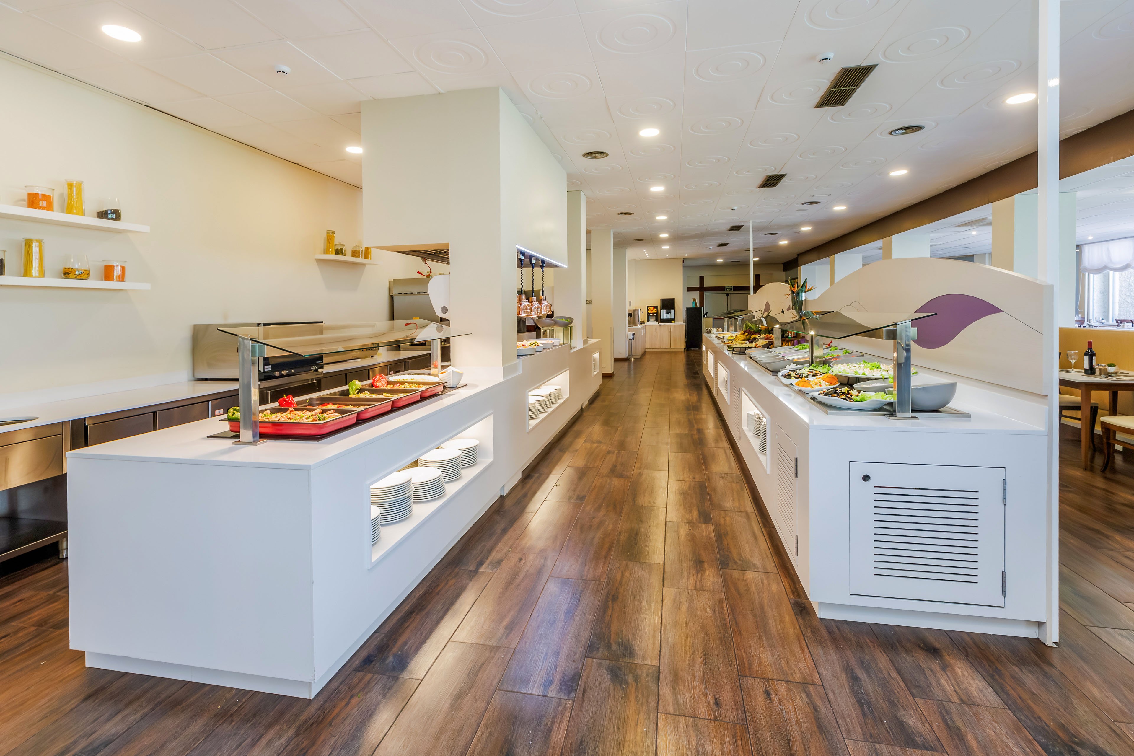 A modern self-service buffet counter is set up in a bright cafeteria or restaurant. Long white serving islands display a variety of dishes and salads under glass sneeze guards. The space features warm wooden flooring, recessed ceiling lights, and clean minimalist decor. No people are present, emphasizing the neat arrangement and contemporary design of the dining area.
