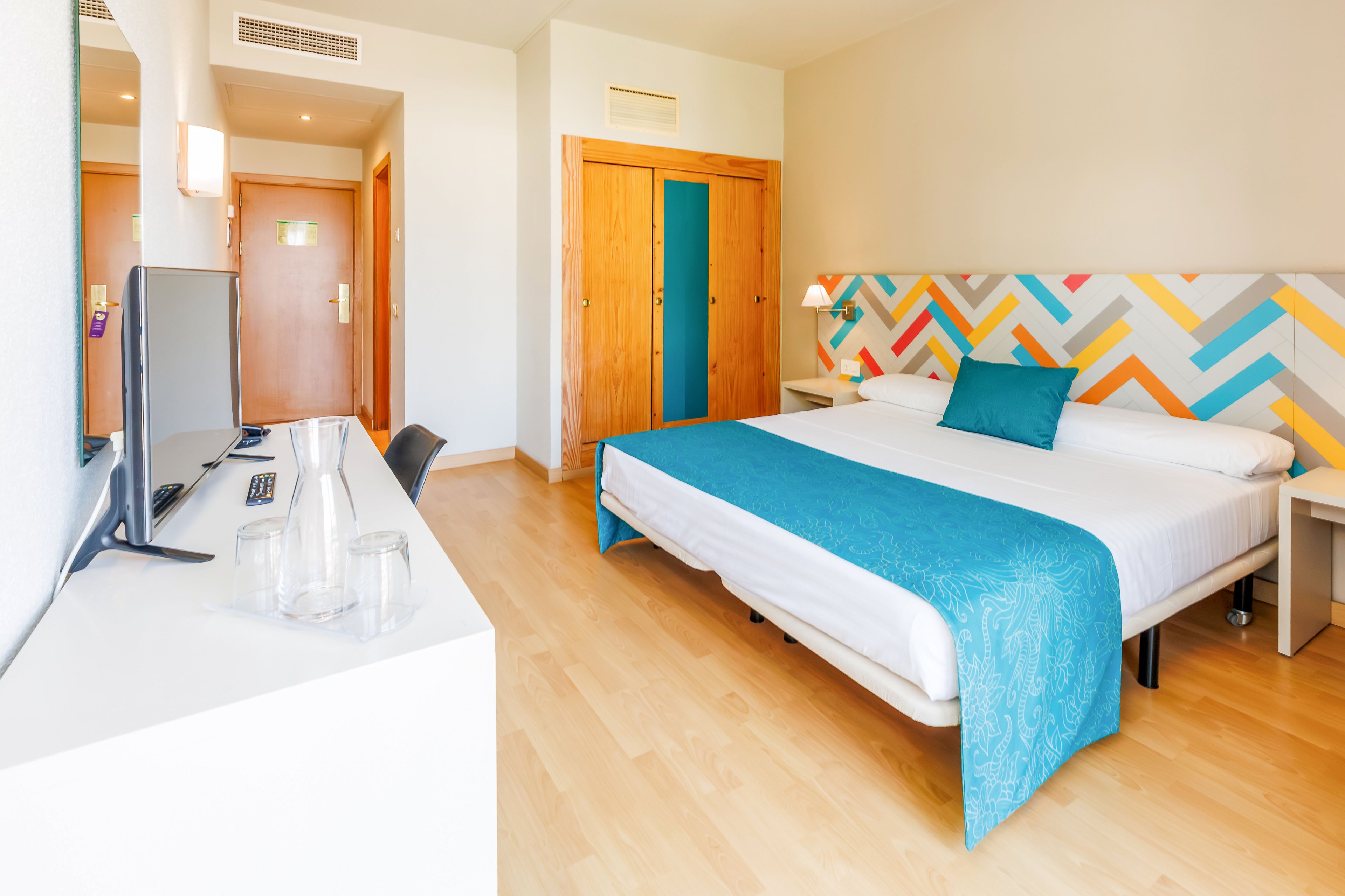 A bright, modern hotel room features a large double bed with white linens and a turquoise bed runner and pillow. The headboard displays a colorful geometric pattern that adds a playful accent to the neutral walls. A flat-screen television sits on a white console opposite the bed, next to clear drinking glasses. Wooden doors and light wood flooring contribute to the warm, inviting atmosphere of the space.