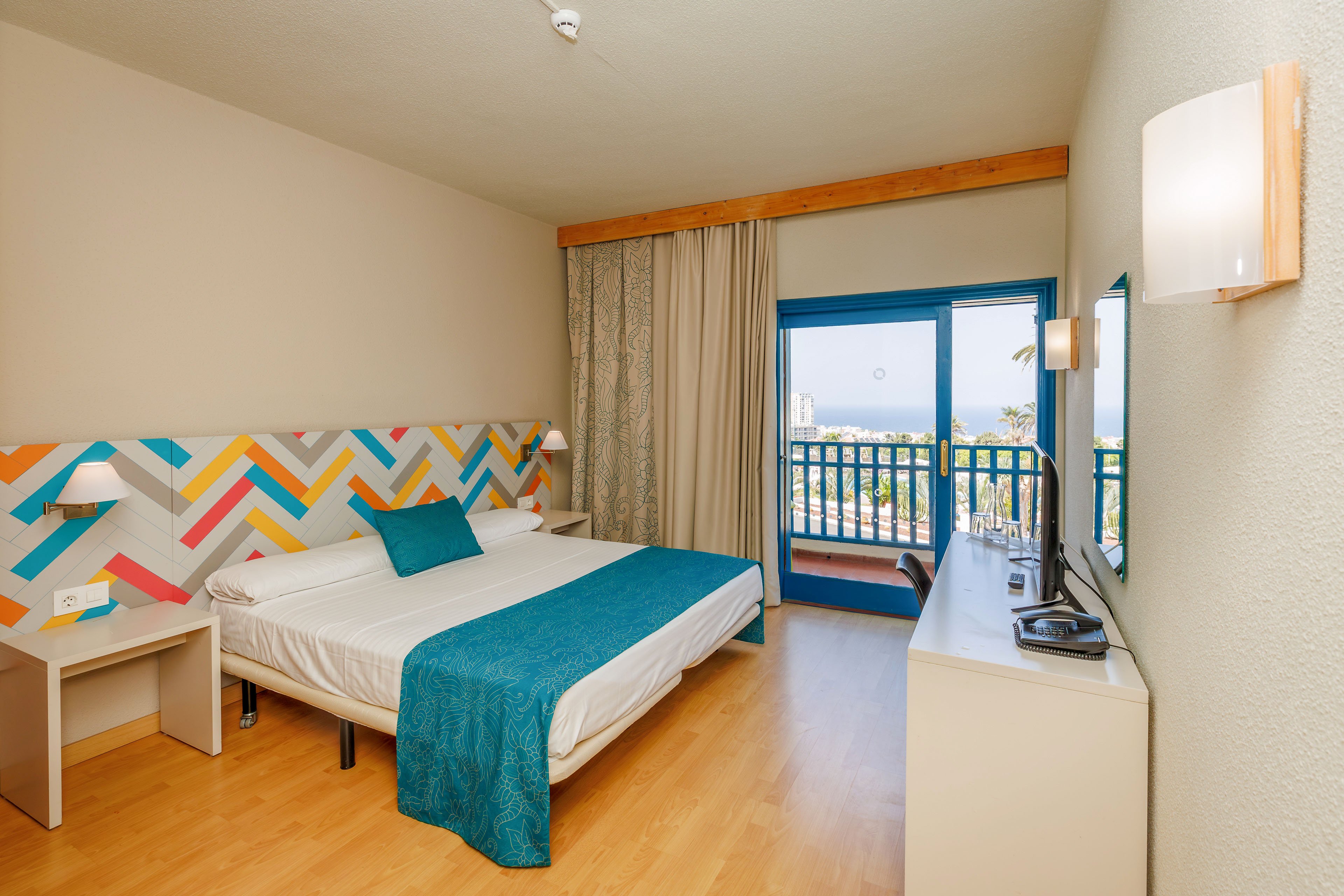 A modern hotel room features a neatly made double bed with white linens and teal accents. The headboard displays a colorful geometric pattern that adds a playful touch to the neutral walls. A large glass door opens to a balcony with a railing, offering an outdoor view and natural light. A desk with a flat-screen television and mirror lines one wall, while wooden flooring and simple lighting create a clean, welcoming atmosphere.