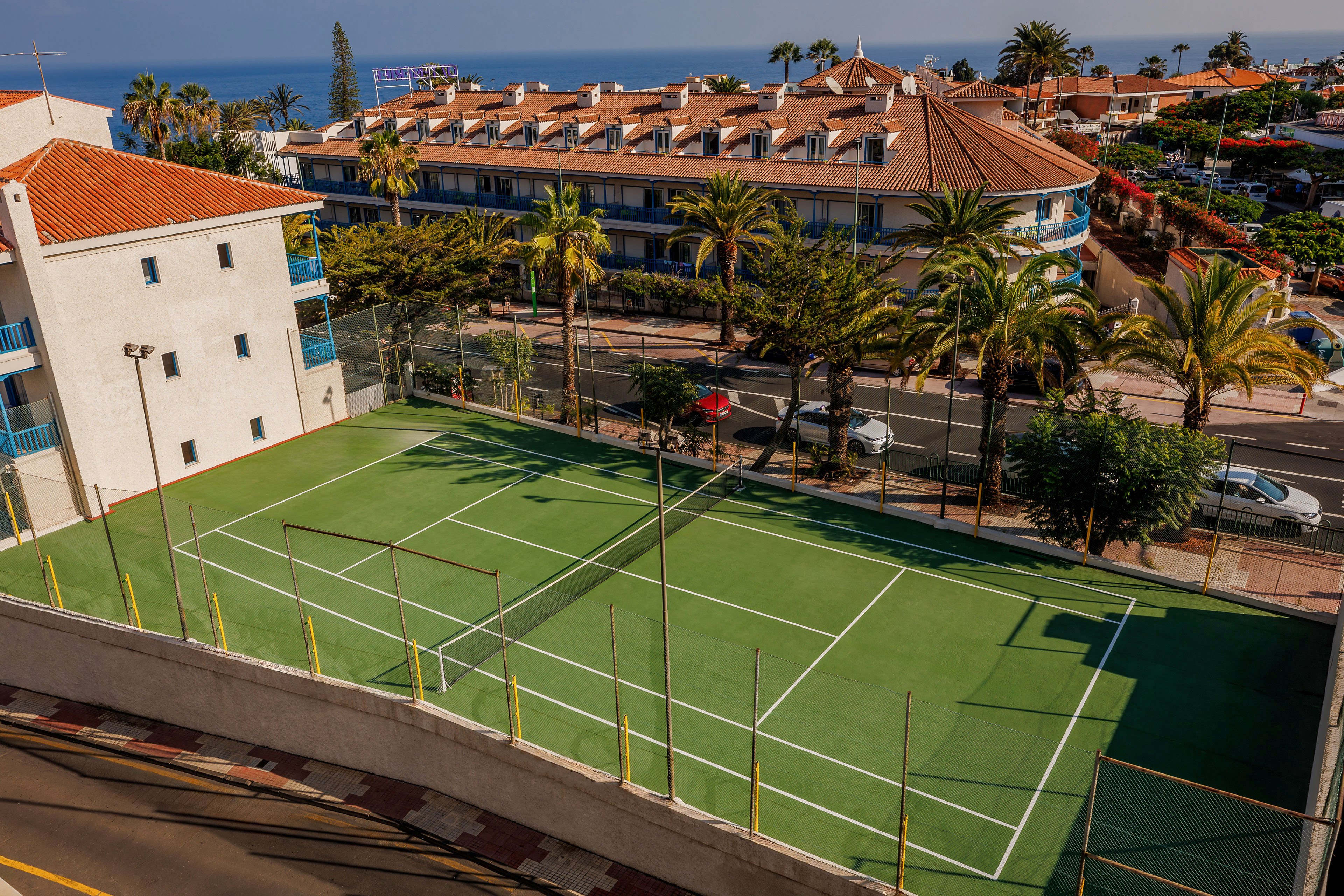 An empty green tennis court is viewed from above in a sunny coastal town. The court is surrounded by high fences and bordered by a street with passing cars and palm trees. Across the road, a large resort-style building with red-tiled roofs and balconies faces the ocean in the distance. Bright daylight and clear skies create a warm, vacation-like atmosphere.