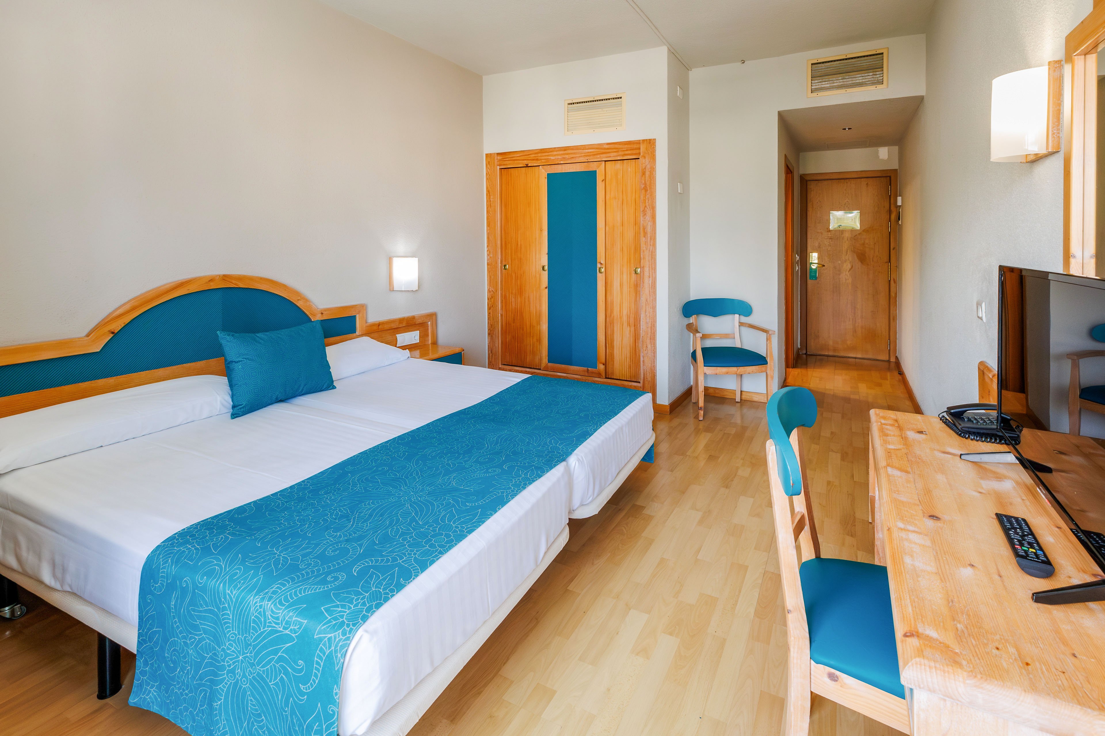 A clean hotel room features two twin beds pushed together with white linens and turquoise runners. The space includes wooden furniture, such as a desk with a flat-screen television and a chair with a turquoise cushion. A wardrobe and a small chair sit near the entrance door, and light wood flooring brightens the room. Soft wall lighting and simple decor create a calm, welcoming atmosphere.
