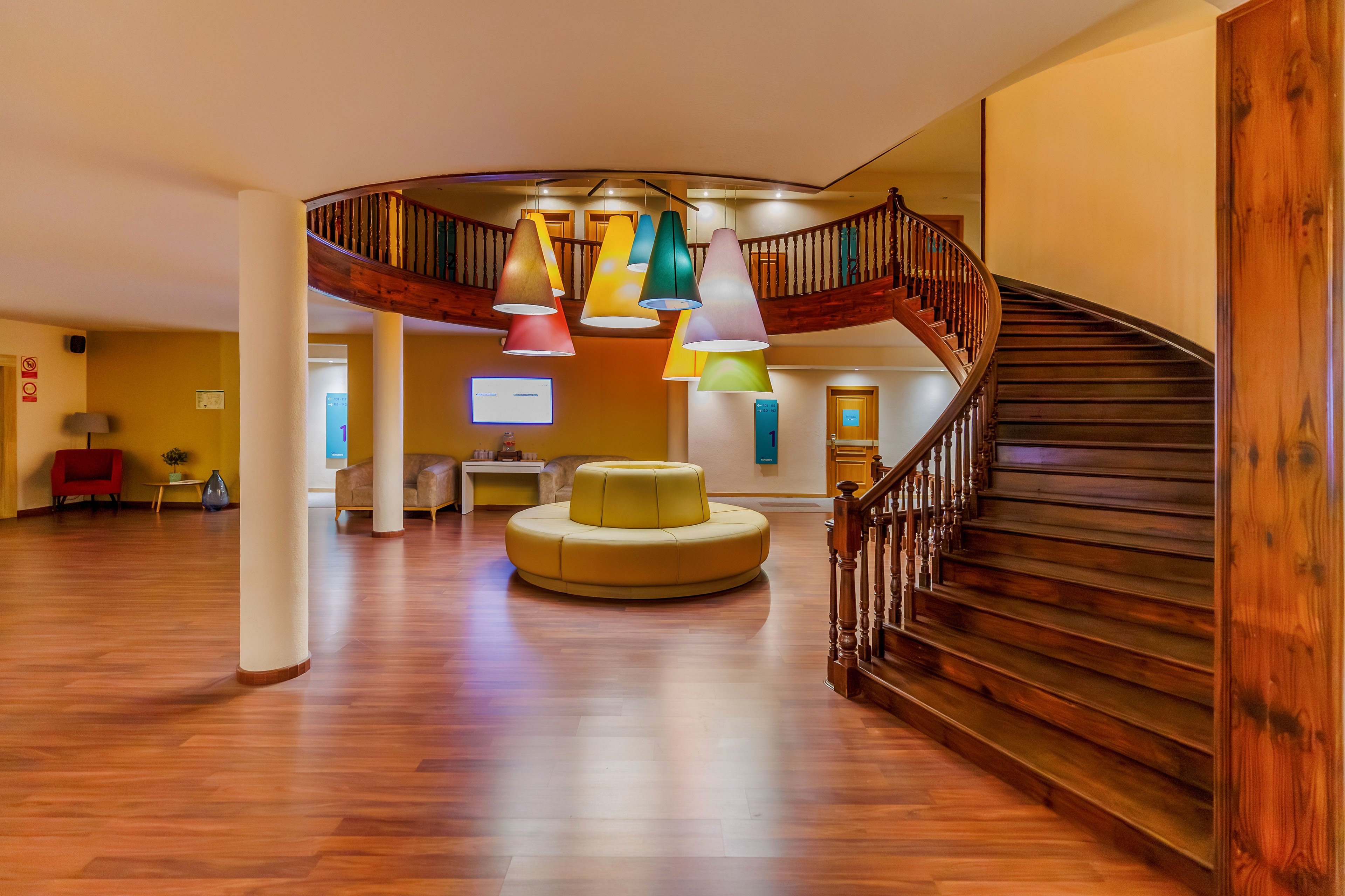 A spacious lobby features a large curved wooden staircase leading to an upper level. In the center of the room, a round upholstered seating area sits beneath a cluster of colorful cone-shaped pendant lights. Warm wood flooring and soft yellow walls create a cozy, inviting atmosphere. A digital screen and small seating area are visible in the background, suggesting a hotel or modern reception space.