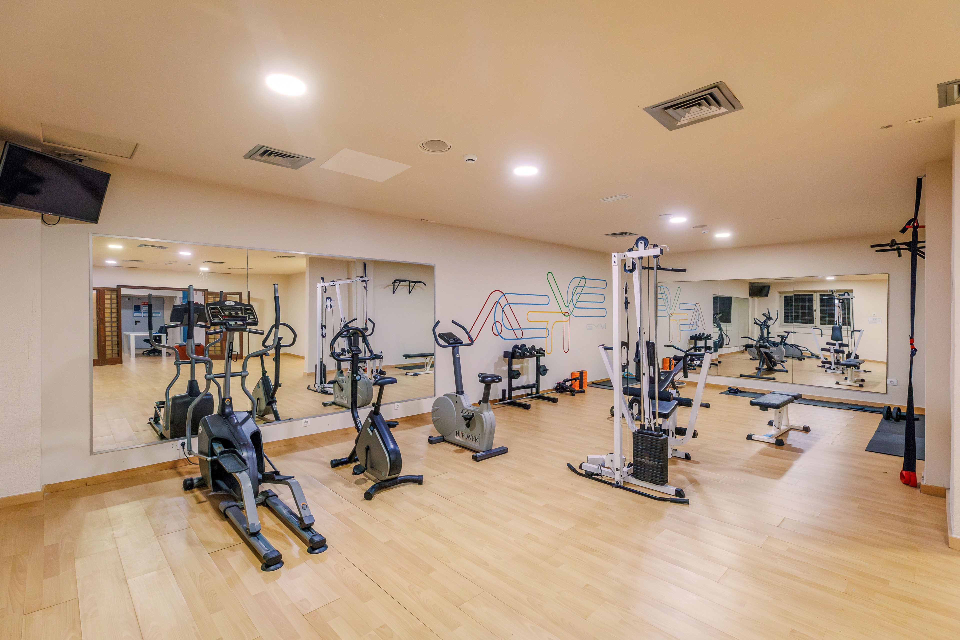 A clean, modern indoor gym features various exercise machines neatly arranged on a light wooden floor. Cardio equipment such as treadmills and stationary bikes stand alongside strength-training machines and free weights. Large wall mirrors visually expand the space and reflect the equipment. Soft, warm lighting and neutral walls create a calm, inviting workout environment.