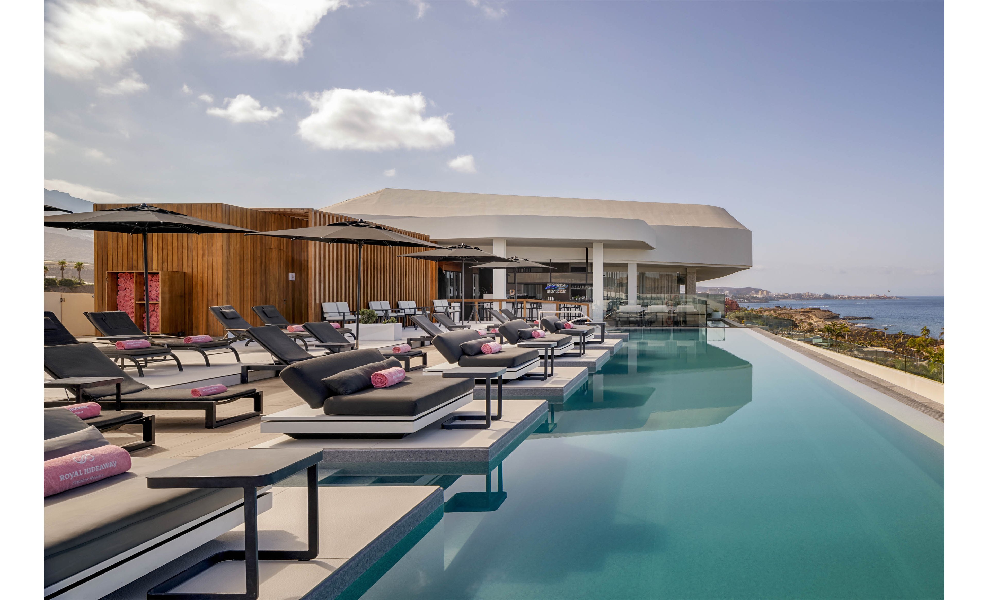 A sleek infinity pool stretches alongside a row of contemporary sun loungers with pink towels. The scene is set on a modern hotel or resort terrace featuring wood and white architectural elements. Calm blue water blends into the distant ocean and rocky shoreline under a partly cloudy sky. The overall atmosphere is luxurious, minimal, and relaxing, suggesting an upscale vacation destination.
