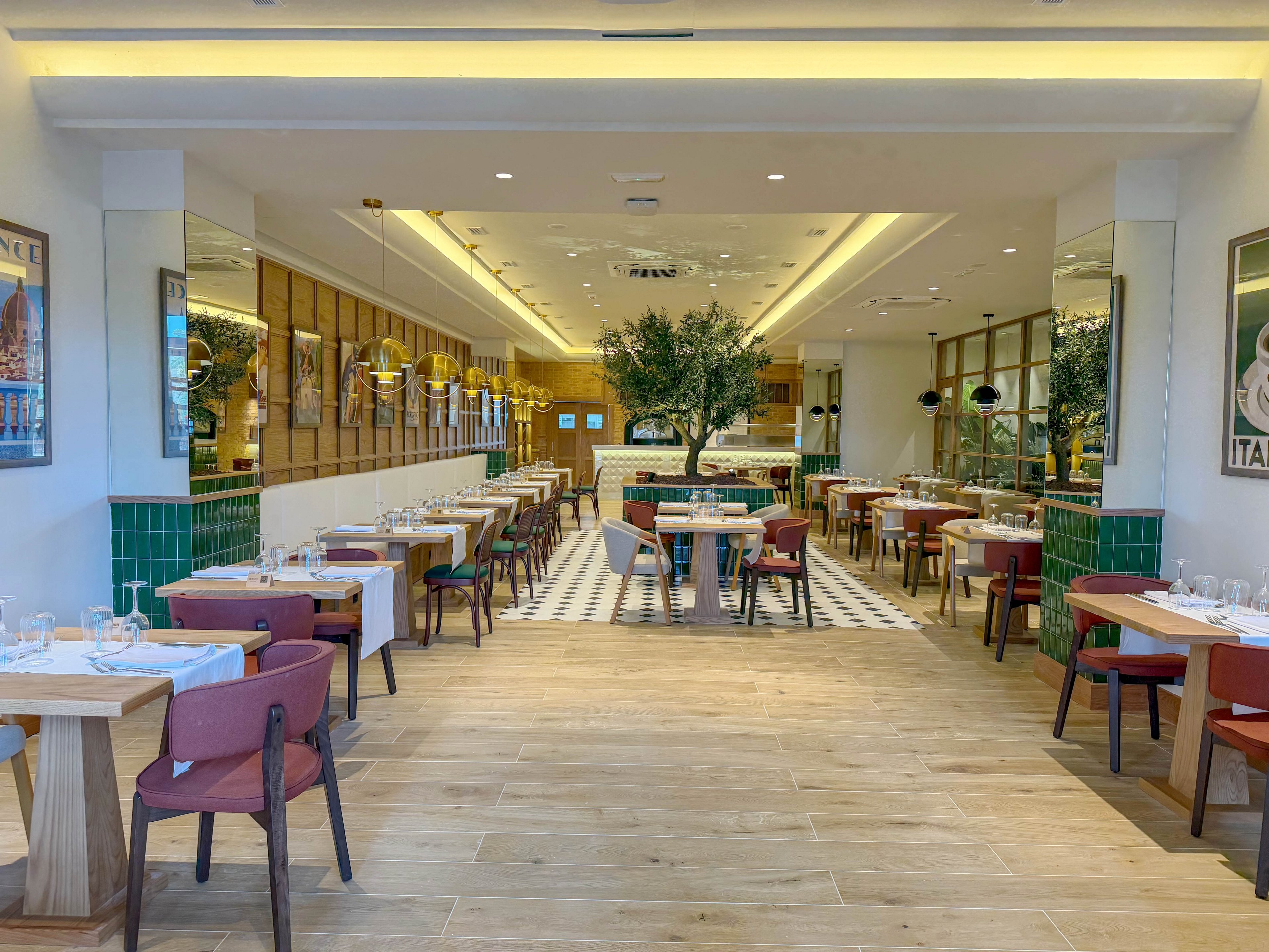 A bright, modern restaurant interior features neatly arranged tables with white tablecloths and wooden chairs. The space is open and symmetrical, with a central aisle leading to a large indoor tree near the back. Warm lighting, green tiled accents, and framed wall art create a welcoming atmosphere. Floor-to-ceiling windows on the right bring in natural light, enhancing the clean and contemporary design.
