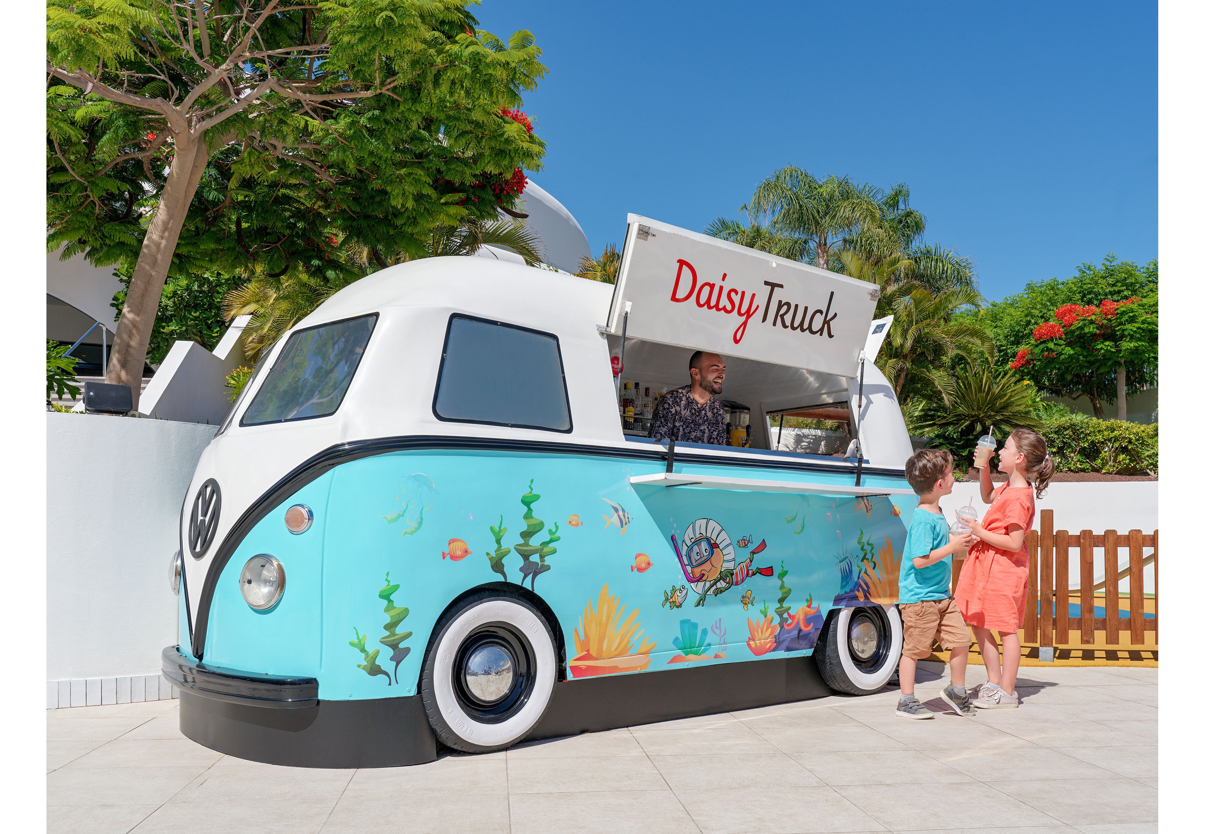 A retro-style food truck named Daisy Truck is parked outdoors under bright blue skies. The vehicle is decorated with colorful underwater illustrations featuring fish, coral, and sea plants. A vendor inside the truck serves two young children standing on the pavement, with tropical trees and a wooden fence in the background. The scene feels cheerful and summery, suggesting a fun vacation or resort setting.