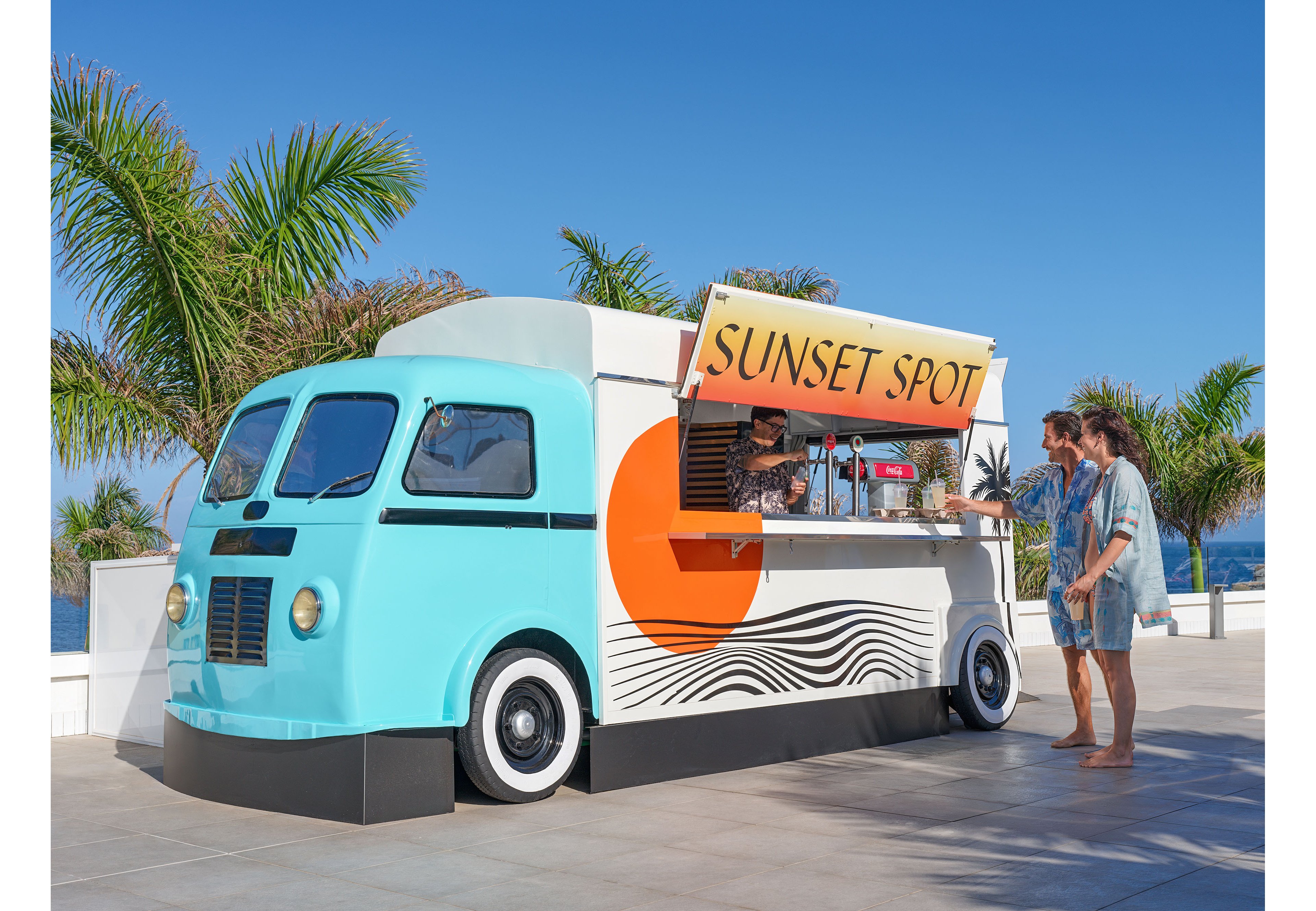 A retro-style food truck with turquoise and white paint is parked on a sunny beachfront promenade. The serving window is open under a sign reading “SUNSET SPOT” as two people order from the vendor inside. Palm trees and a clear blue sky frame the relaxed coastal setting. Bold graphic shapes and waves decorate the truck, enhancing the vibrant, vacation atmosphere.