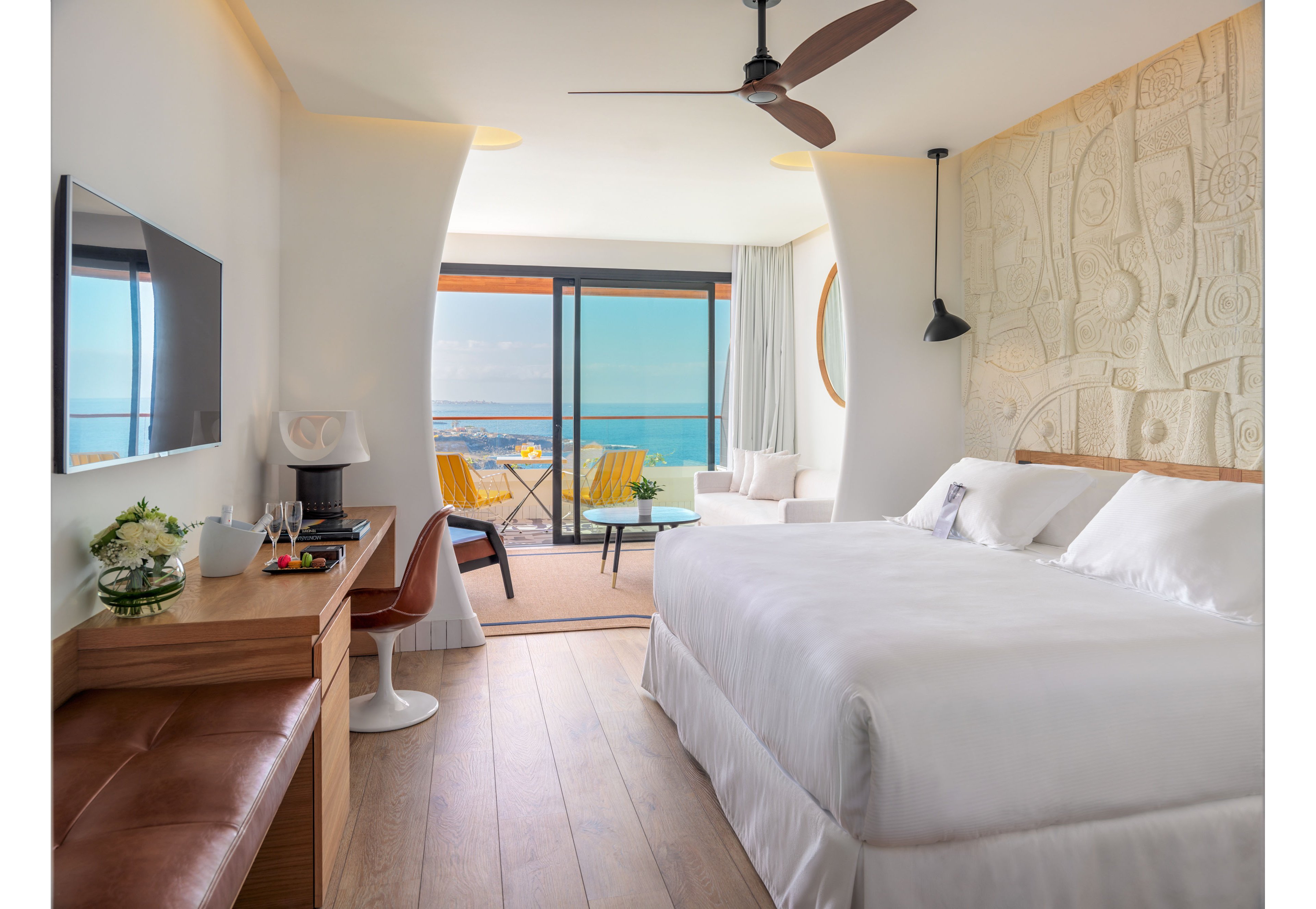 Bright contemporary hotel room featuring a large bed with white linens and textured wall art. A wooden desk with a television, chair, and refreshments sits along one side, opposite a leather bench. Floor-to-ceiling glass doors open to a balcony with yellow chairs and a table overlooking the ocean and coastline. Natural light, soft neutral tones, and minimalist decor create a calm, inviting atmosphere.