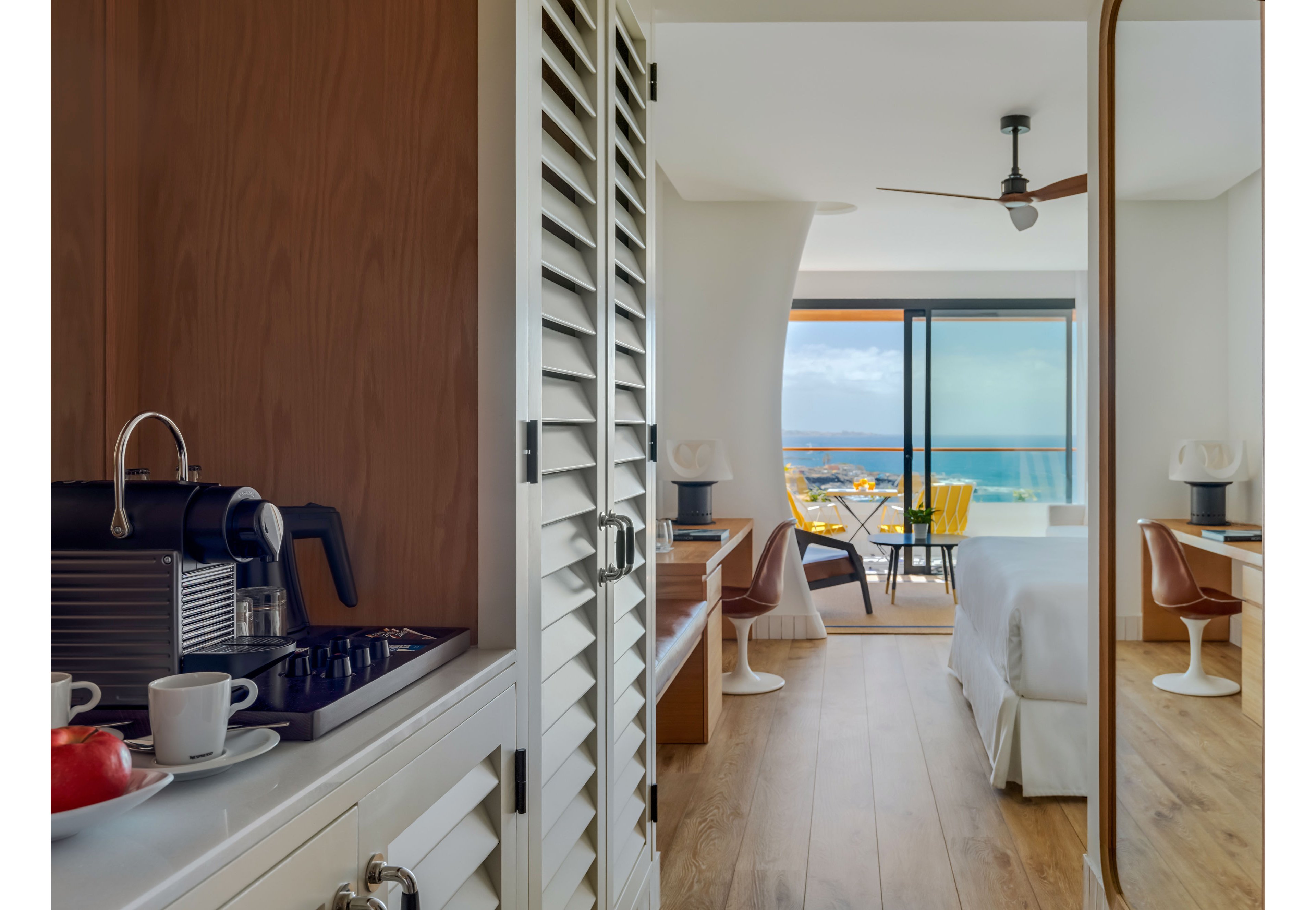 A contemporary hotel room features a cozy bed and wooden flooring leading to a balcony overlooking the ocean. In the foreground, a small kitchenette with a coffee machine, cups, and fruit adds convenience for guests. The room includes a desk area with a chair and lamp, and large sliding glass doors fill the space with natural light. Outside, yellow chairs and a table invite relaxation while enjoying the coastal scenery.