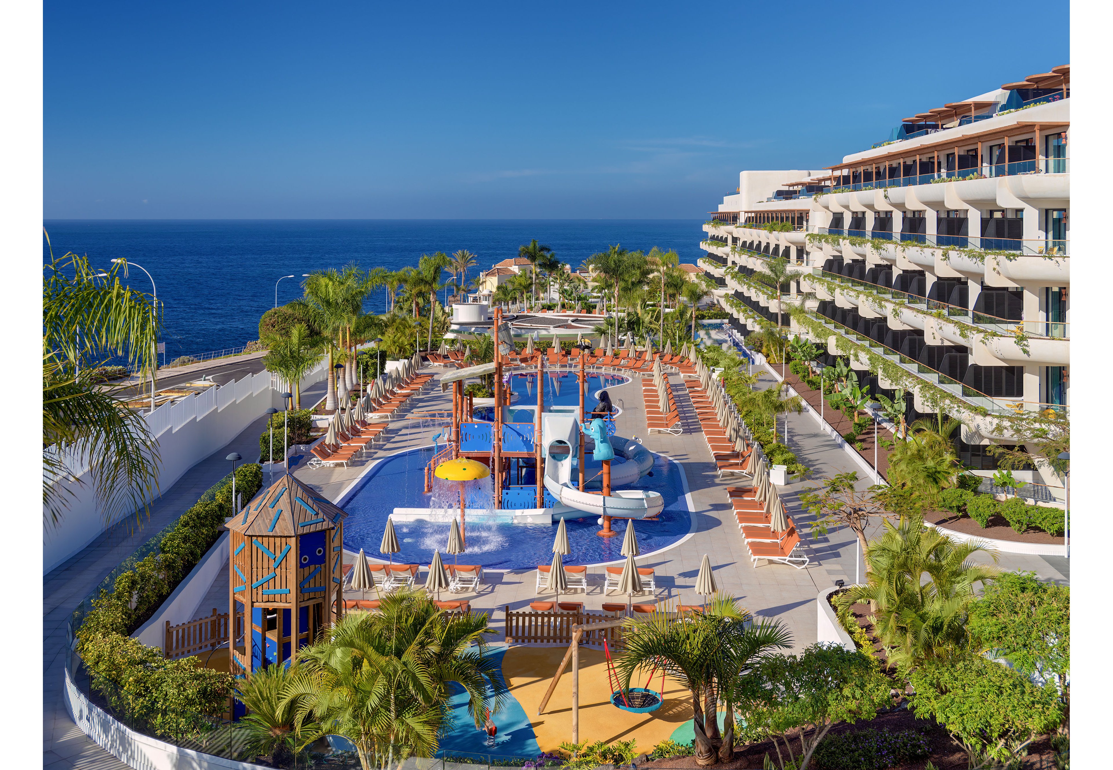 A large coastal resort features an expansive outdoor pool and colorful water playground overlooking the ocean. Sun loungers and umbrellas line the poolside terraces, with palm trees and lush greenery adding a tropical feel. The water park includes slides, splash structures, and a small play tower designed for children. Multi-story hotel buildings with balconies rise along one side, framing the bright blue sky and sea in the background.