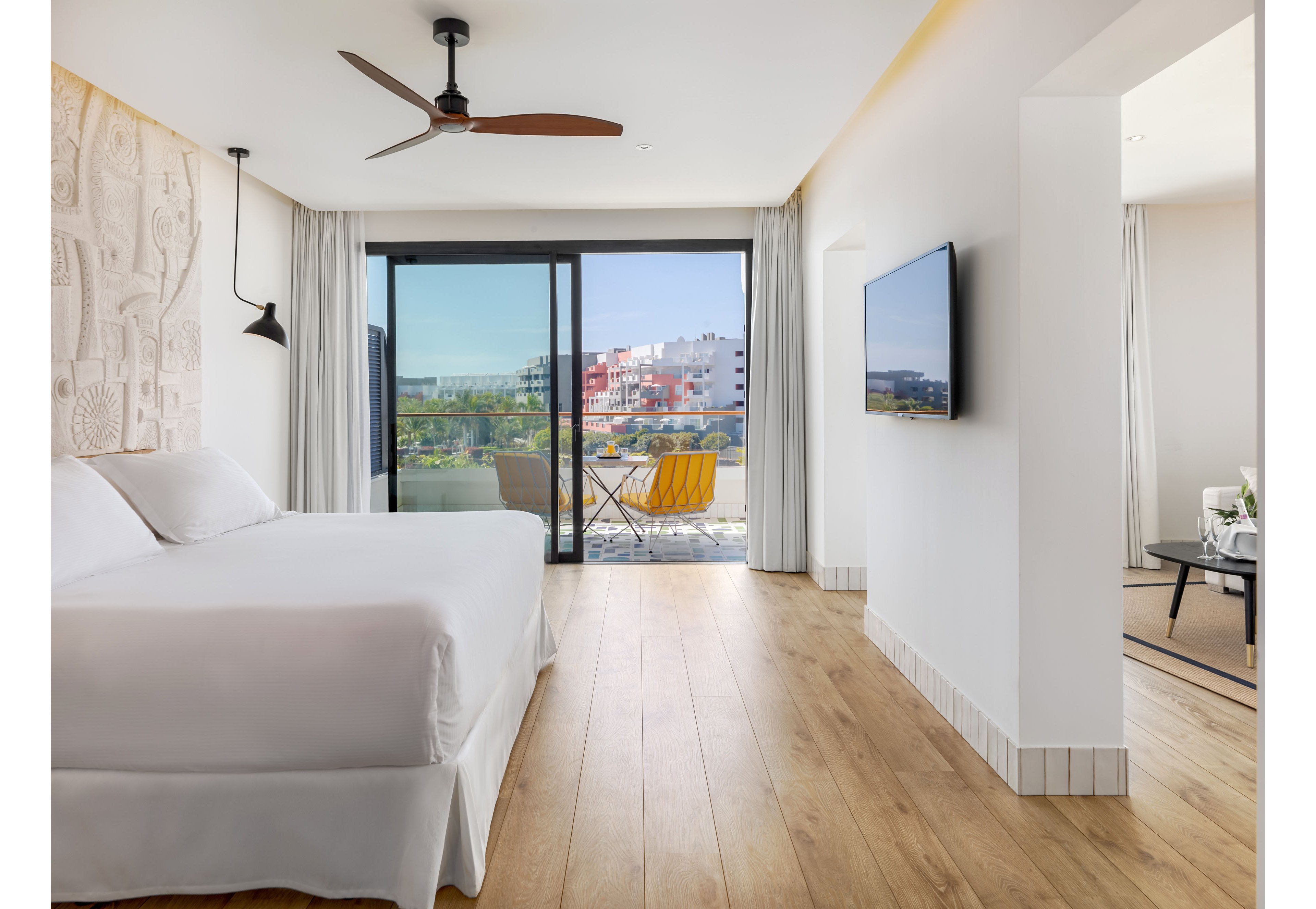Bright contemporary hotel room featuring a large neatly made bed and light wood flooring. A sliding glass door opens to a balcony with yellow chairs and a view of colorful buildings and greenery outside. The interior includes white walls, a ceiling fan, wall-mounted television, and minimalist decor. An adjacent sitting area with a small table and chairs is visible through an open doorway, enhancing the spacious feel.