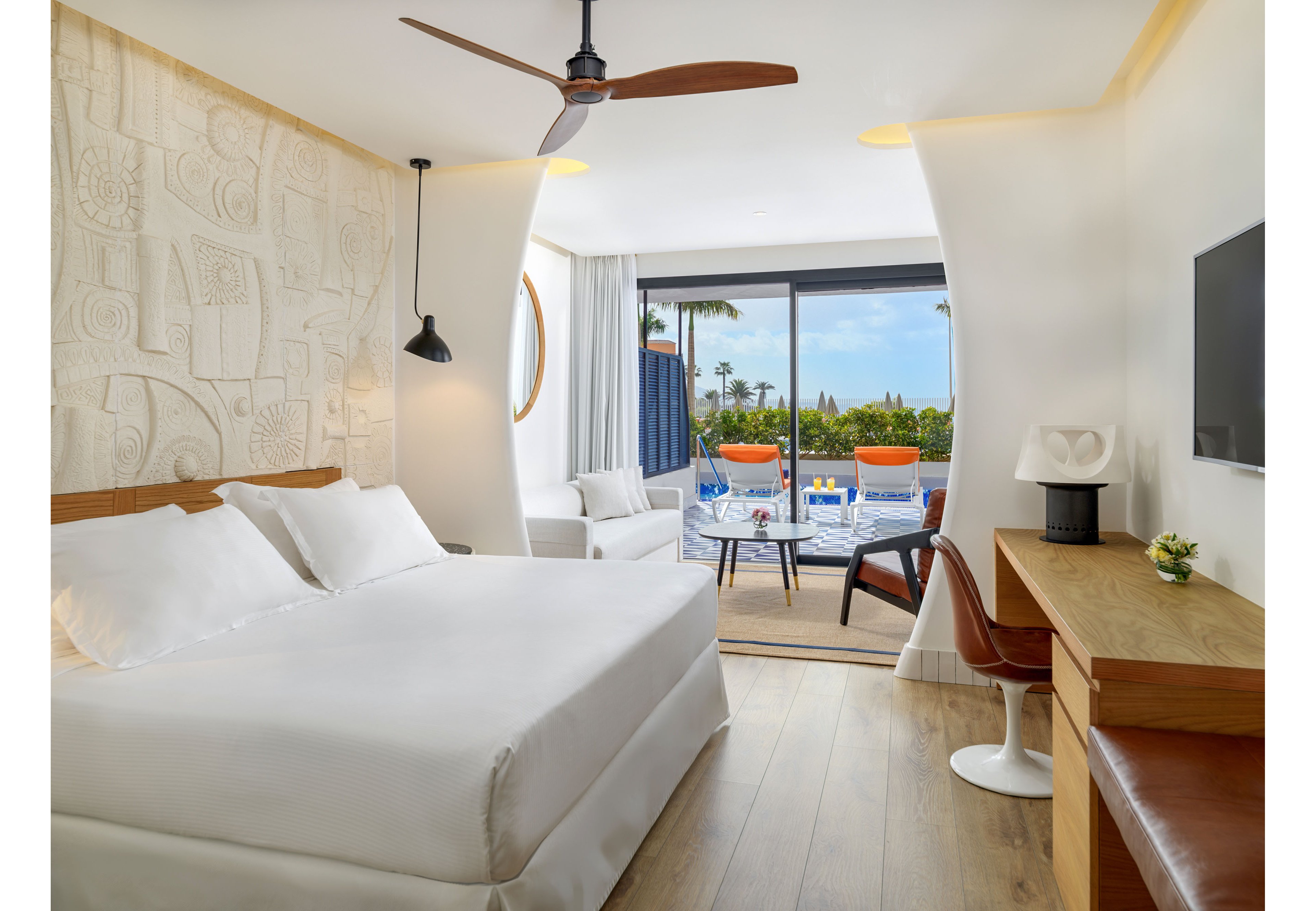 A contemporary hotel room features a large bed with white linens and a textured artistic wall. A wooden desk with a chair and a mounted television sit opposite the bed, while a ceiling fan hangs above. Floor-to-ceiling glass doors open to a terrace with lounge chairs and a view of a pool and lush greenery. Natural light fills the space, creating an airy and inviting atmosphere.