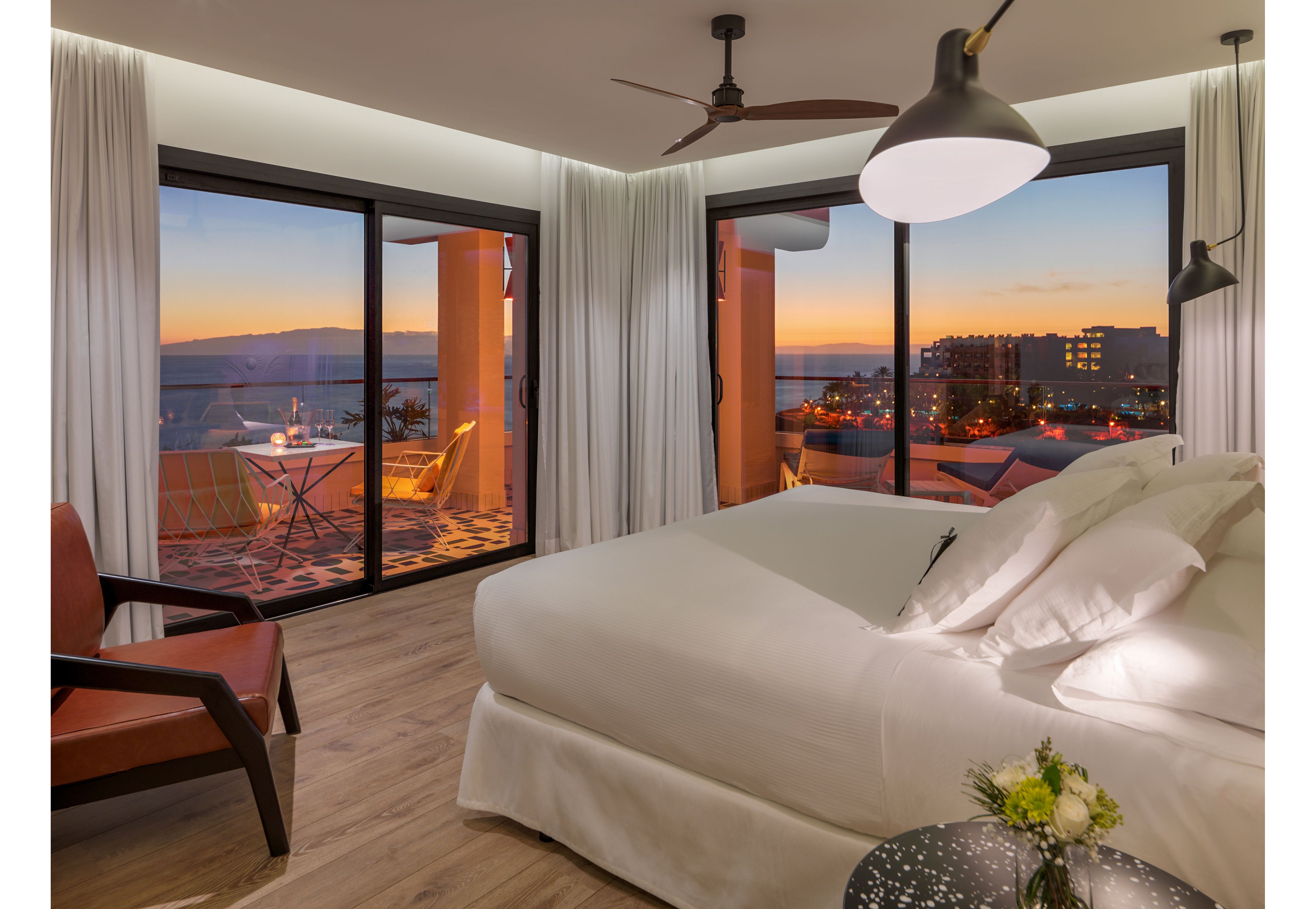 A contemporary hotel bedroom features a large bed with white linens and a leather armchair on wooden flooring. Floor-to-ceiling windows open to a balcony with a small table and chairs overlooking a coastal city. The warm sunset sky casts orange and purple hues over the ocean and surrounding buildings. Soft interior lighting and minimalist decor create a calm, inviting atmosphere.