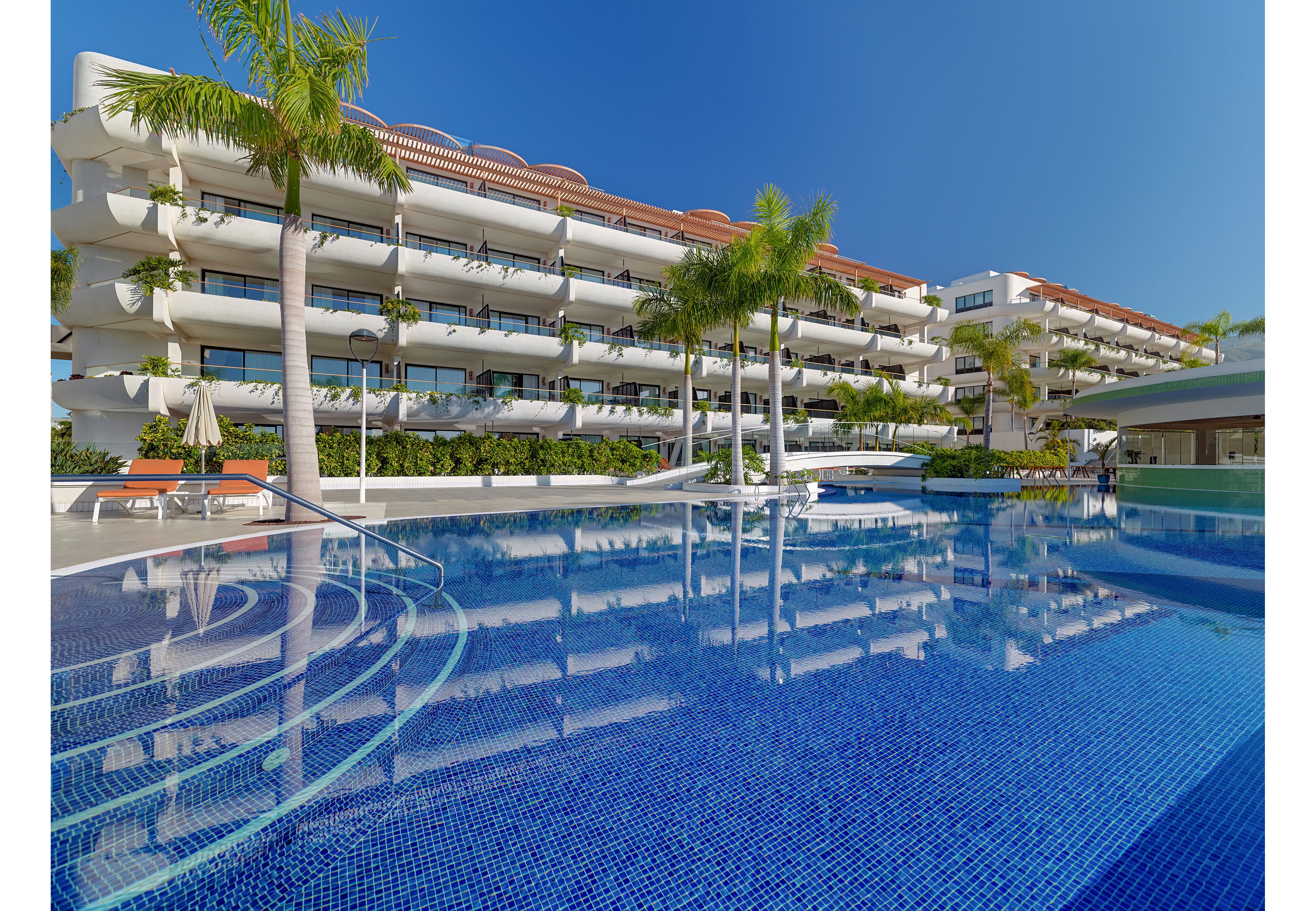 A contemporary multi-story resort hotel overlooks a large outdoor swimming pool under a clear blue sky. Palm trees and lush greenery surround the pool area, adding a tropical atmosphere. Sun loungers and shaded seating are visible along the poolside, suggesting a relaxing vacation setting. The calm water reflects the building’s balconies and architectural details, emphasizing the clean and modern design.