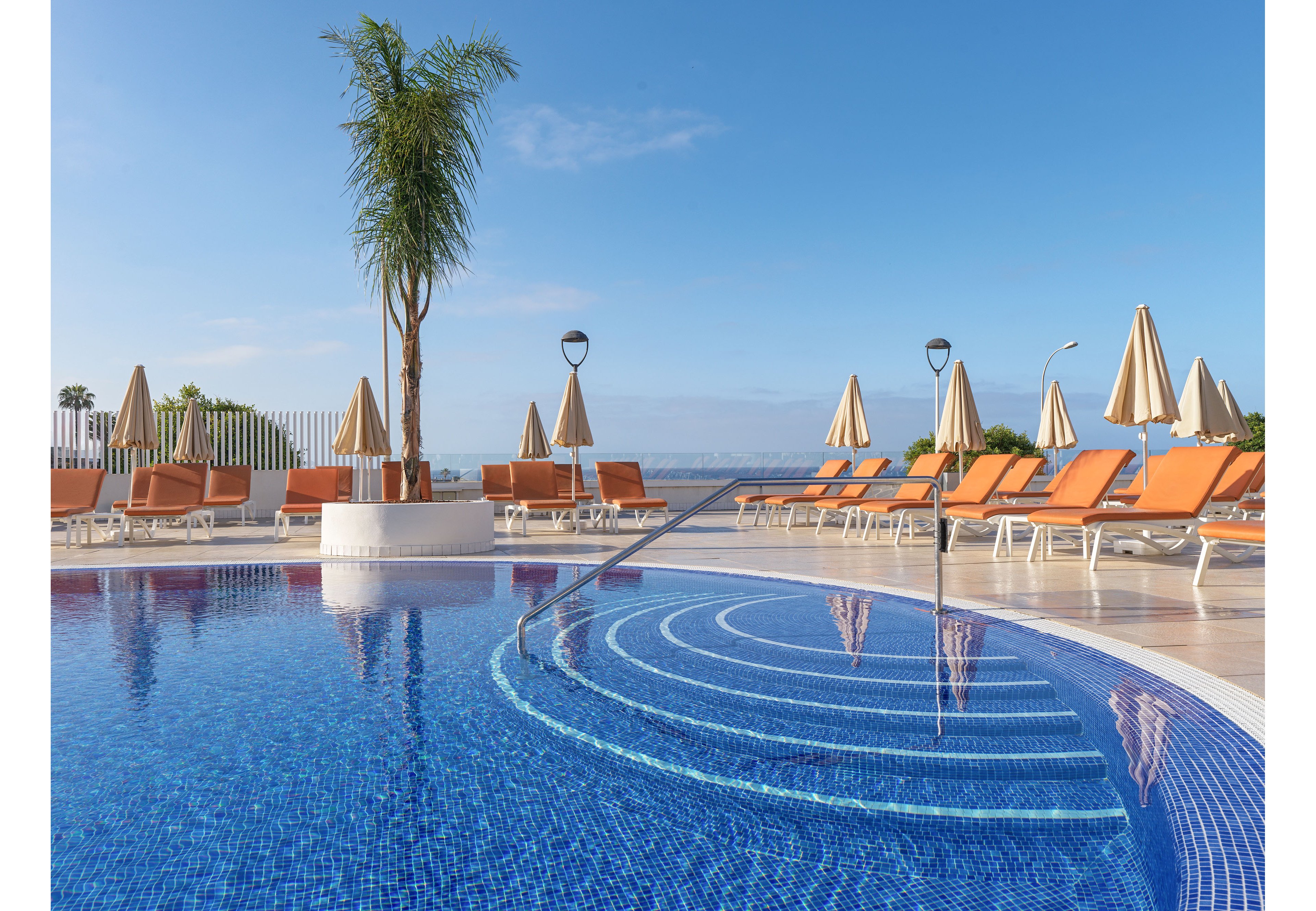 A curved outdoor swimming pool with blue mosaic tiles sits in the foreground under a clear blue sky. Orange sun loungers and beige umbrellas line the poolside terrace, creating a relaxed vacation atmosphere. A single palm tree rises from a white planter near the pool steps, adding a tropical touch. The scene overlooks a distant waterfront, suggesting a coastal resort setting.