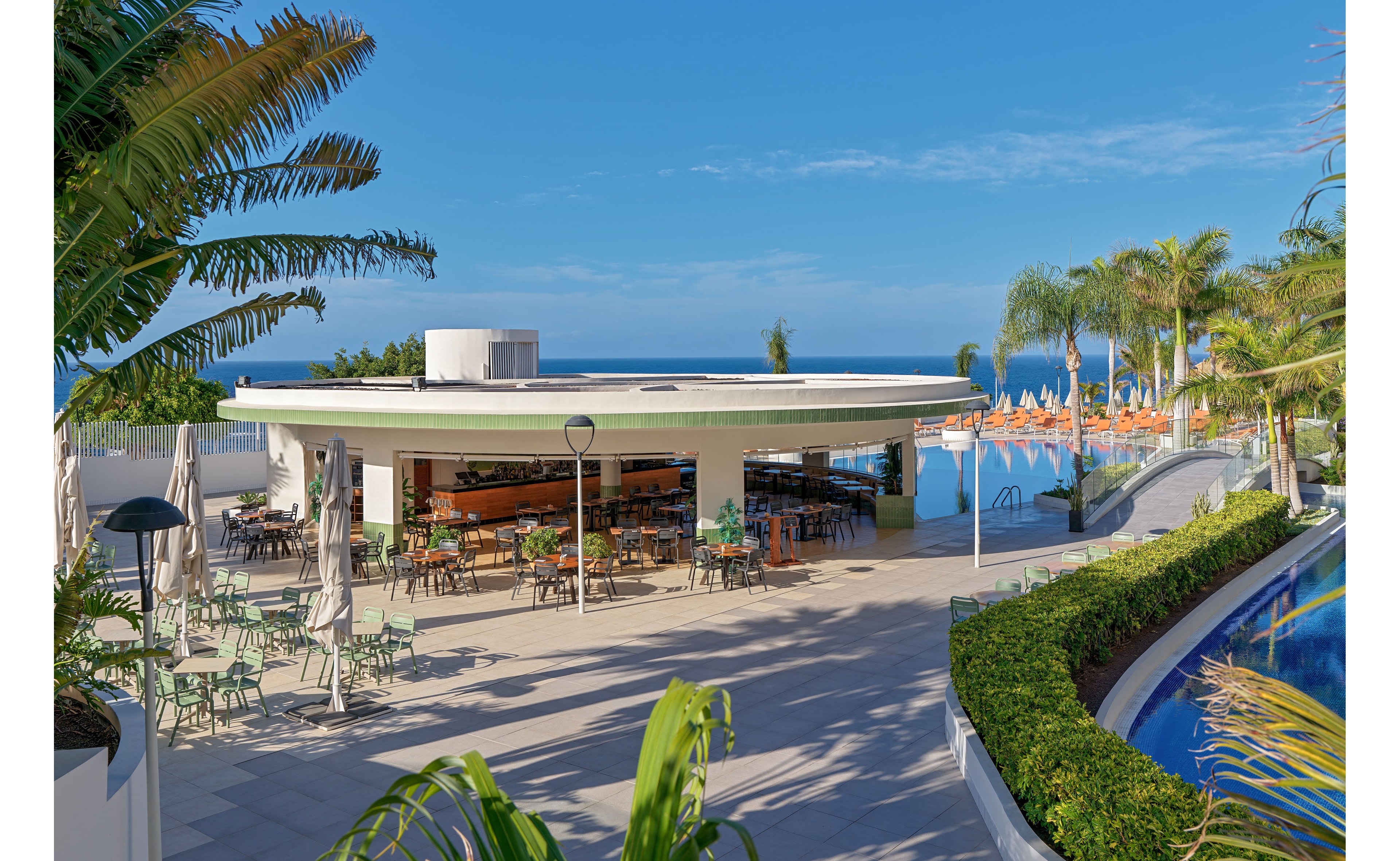 An open-air circular bar sits beside a curved swimming pool at a modern seaside resort. Tables, chairs, and sun umbrellas are arranged on the terrace, with rows of orange sun loungers visible in the background. Lush tropical plants and palm trees frame the scene under a clear blue sky. The ocean can be seen in the distance, enhancing the relaxed vacation atmosphere.