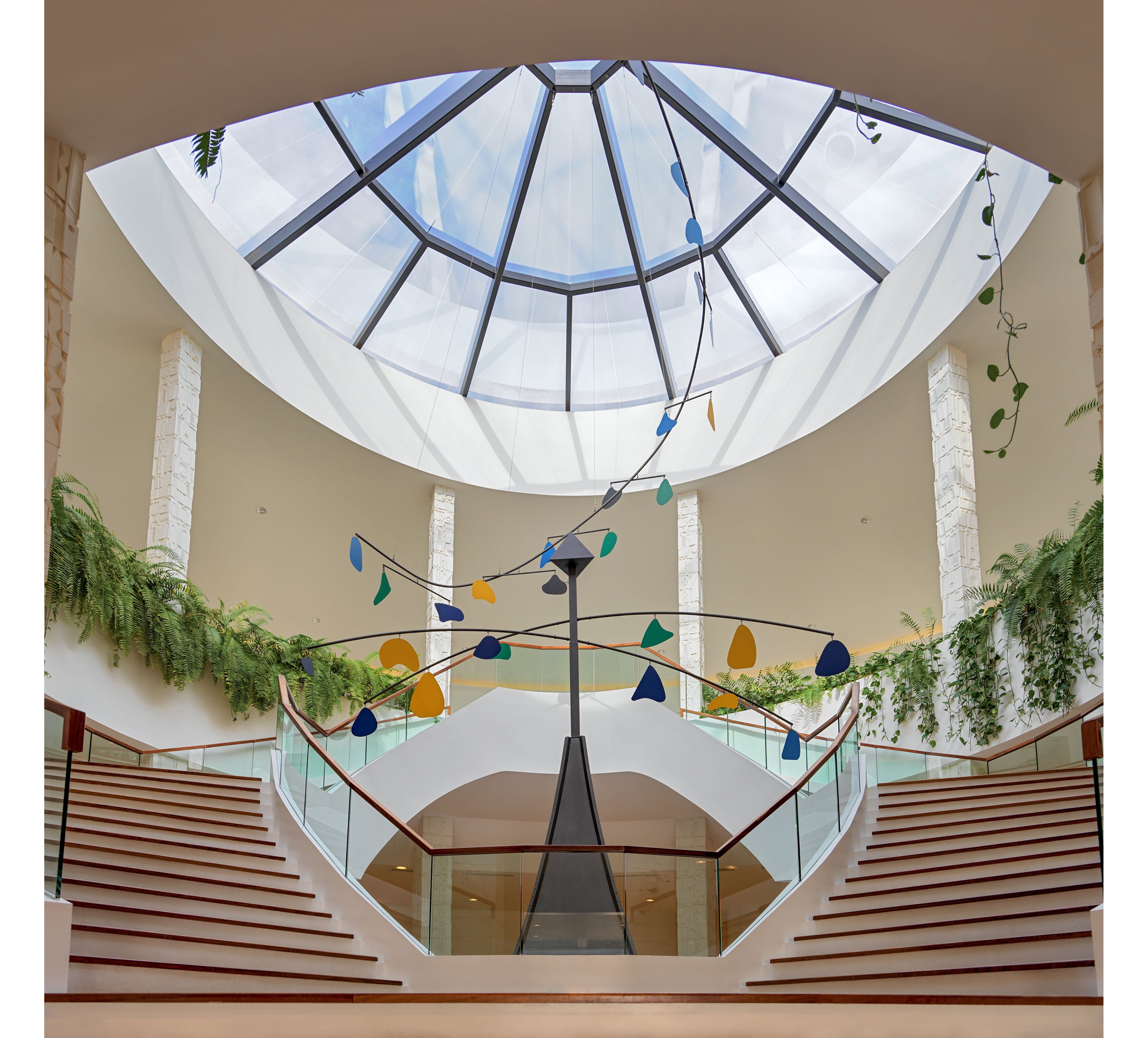 A spacious indoor atrium features a large geometric glass skylight overhead. A colorful hanging mobile with abstract shapes is suspended above a central staircase. Twin curved stairways with glass railings lead down to a lower level, framed by stone columns. Lush green plants cascade along the upper balcony, adding natural contrast to the clean architectural lines.