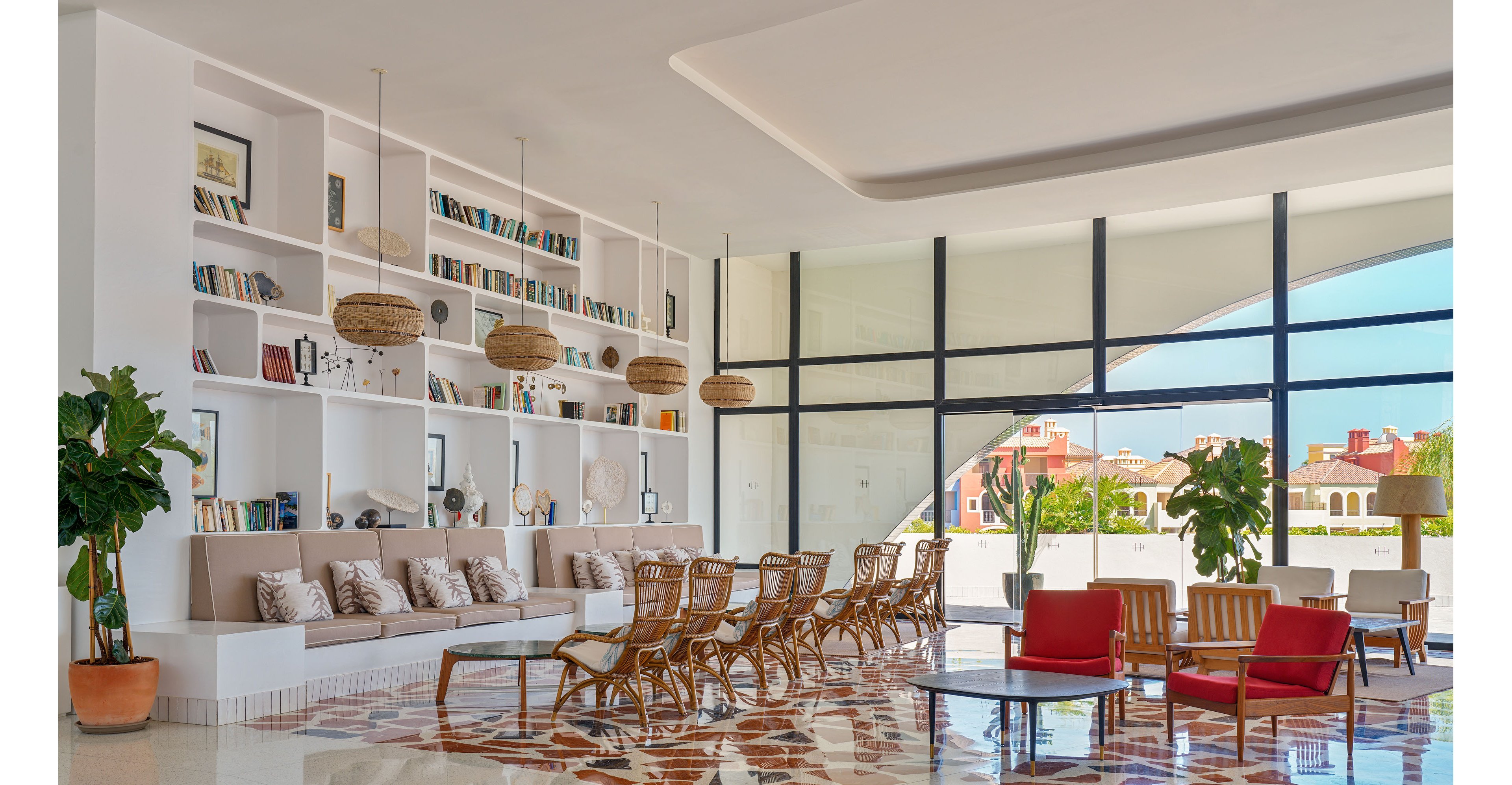 A spacious, contemporary lobby features a long built-in sofa lined with patterned cushions and wicker lounge chairs. Floor-to-ceiling windows flood the room with natural light and reveal colorful buildings outside. White shelving walls are filled with books and decor objects, while woven pendant lights hang overhead. The polished floor with geometric tiles adds warmth and character to the airy interior.