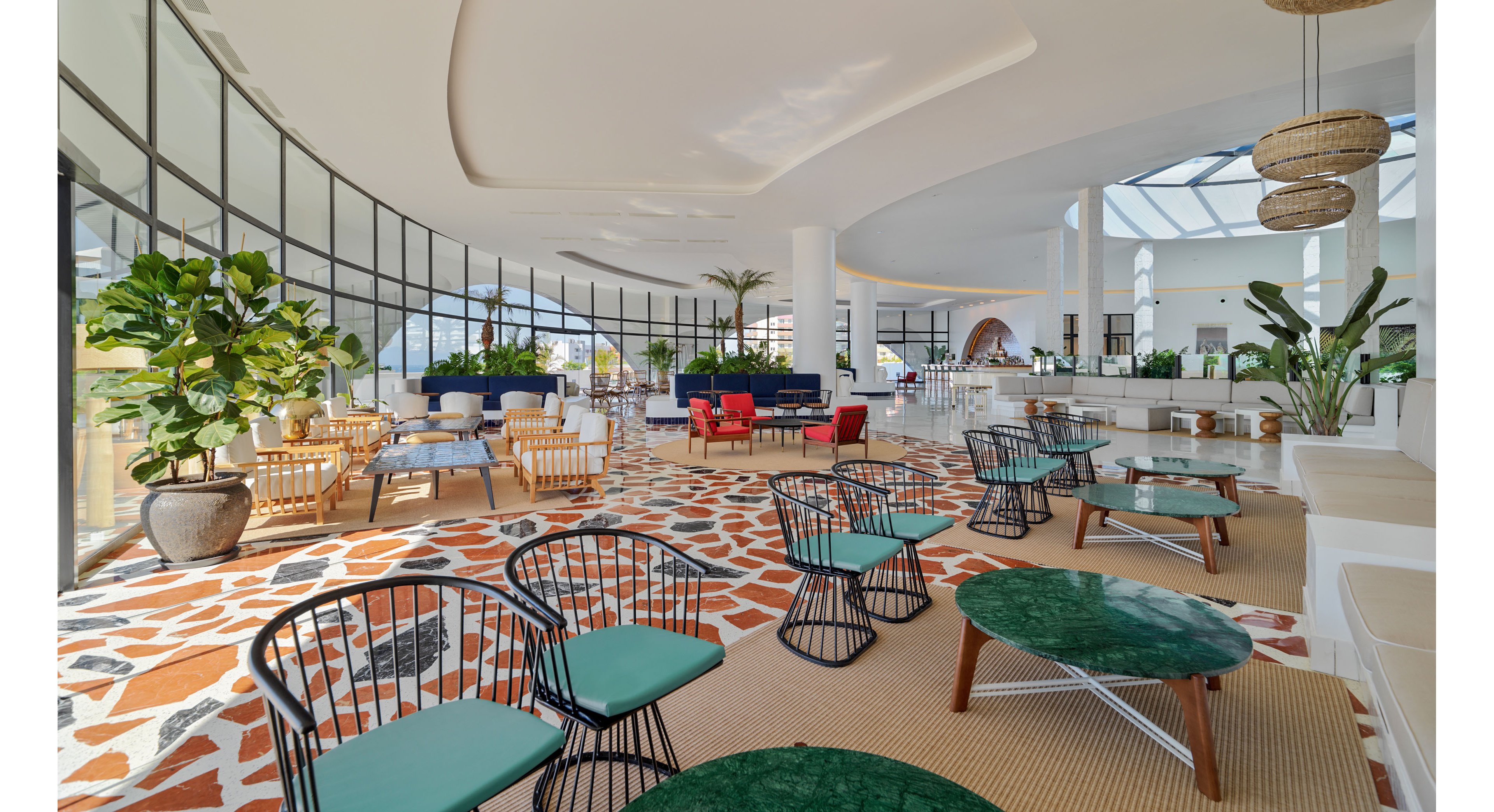 Spacious modern lounge area features a mix of stylish chairs and low tables arranged across a patterned floor. Large floor-to-ceiling windows curve along one side, filling the space with natural light and views outside. Green plants, woven pendant lights, and colorful seating groups create a relaxed yet upscale atmosphere. The open layout leads toward a bar or service counter in the background, enhancing the social and welcoming feel.