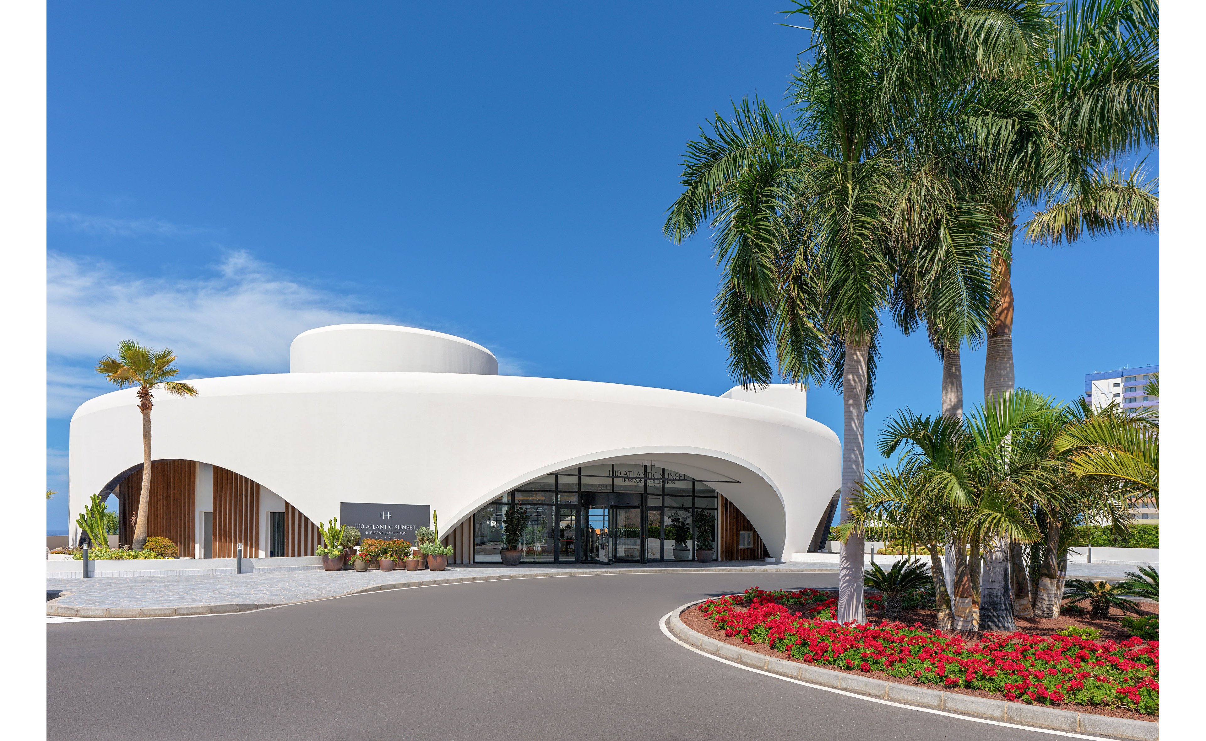 A contemporary white building with smooth, curved architecture stands beside a wide paved driveway. Tall palm trees and vibrant red flower beds frame the entrance, adding lush greenery to the scene. Large arched glass windows reveal the interior while reflecting the bright blue sky. The overall setting appears clean, sunny, and inviting, suggesting a modern public or commercial space.