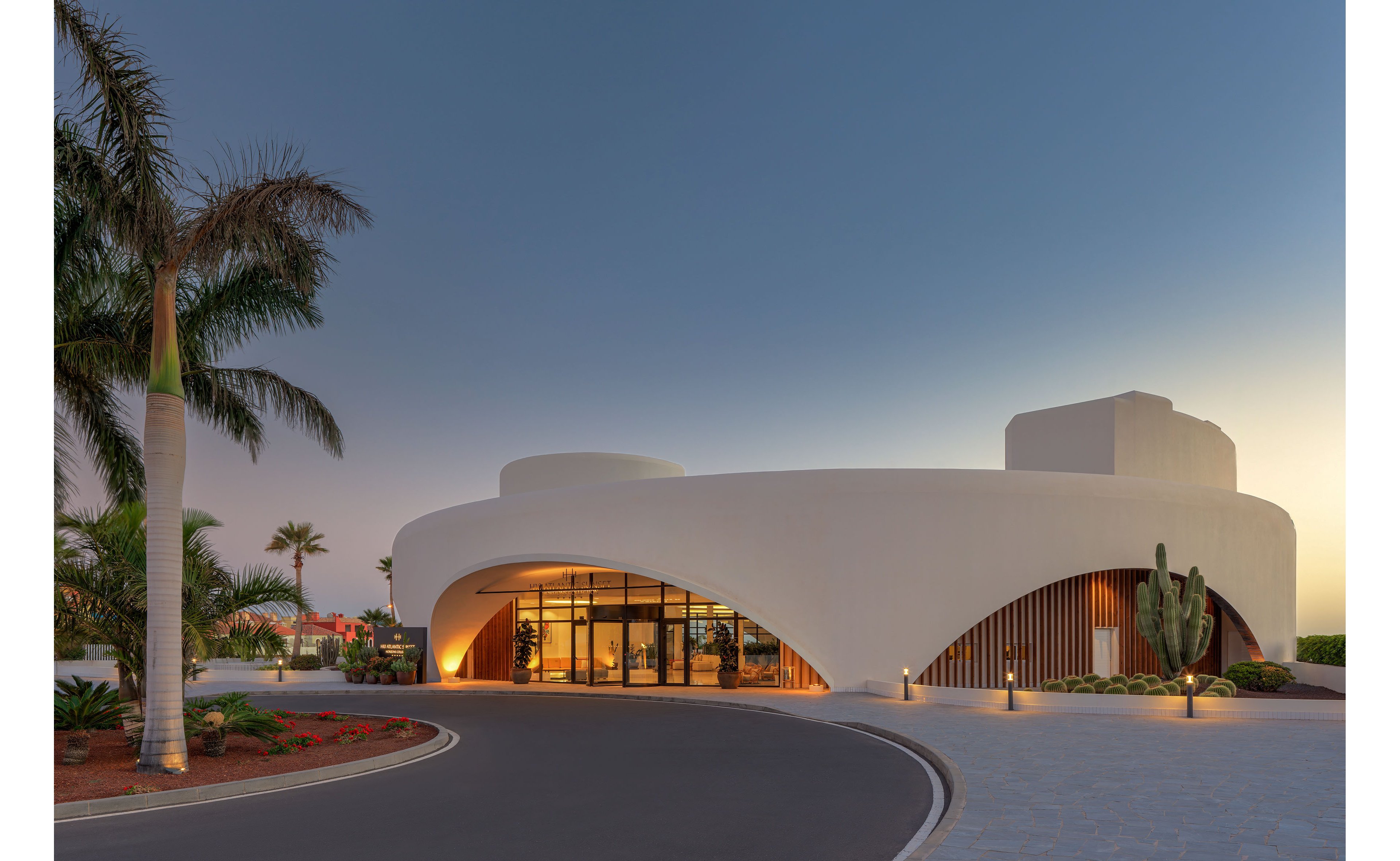 A contemporary white building with smooth, curved architecture stands beside a circular driveway at dusk. Large glass doors and windows reveal a warmly lit interior, contrasting with the cool evening sky. Palm trees, cacti, and landscaped flower beds surround the structure, adding a tropical feel to the scene. Soft ground lights accentuate the entrance and pathways, highlighting the building’s modern design.