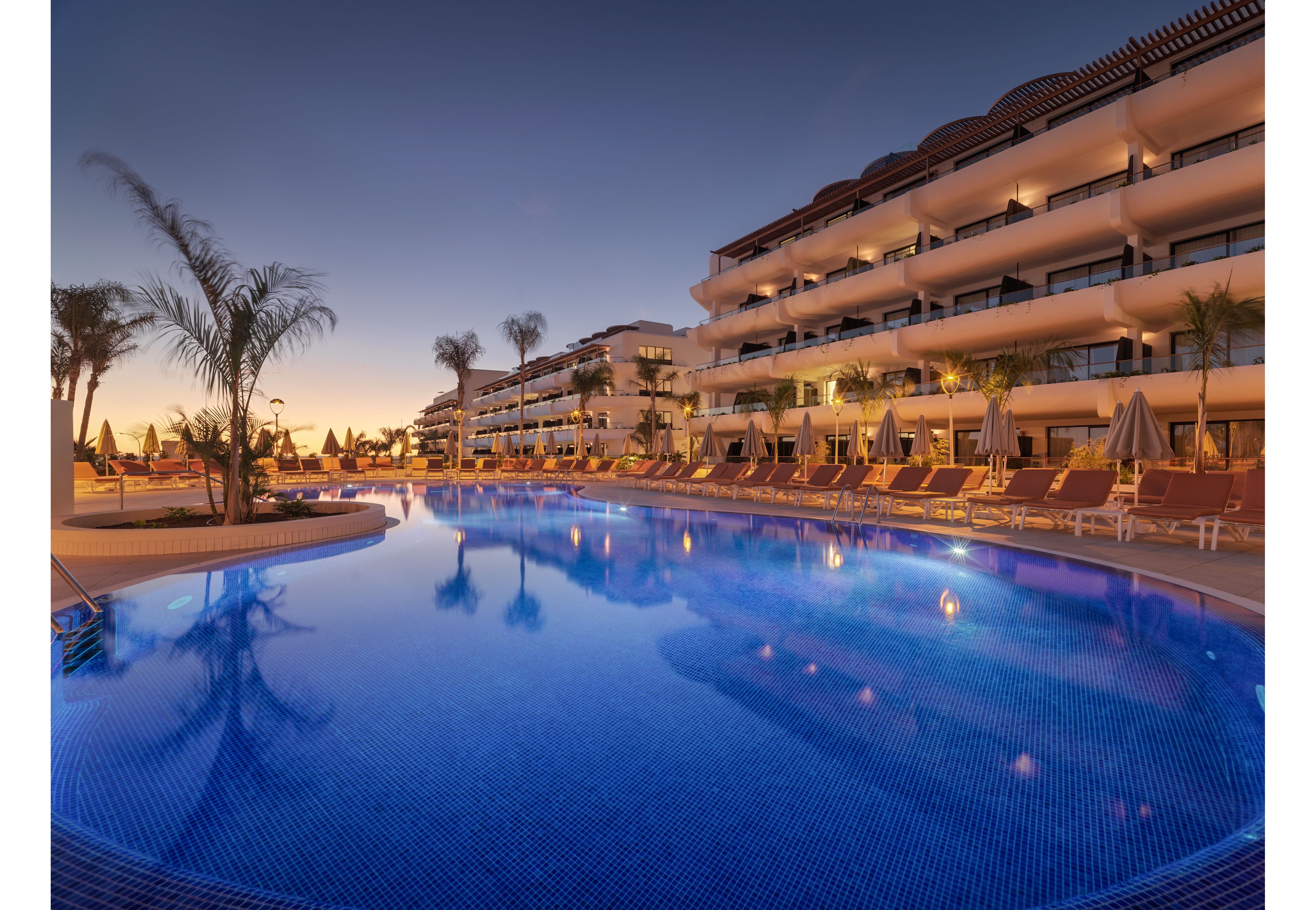 A large, curved swimming pool dominates the foreground of a modern resort at dusk. The calm blue water reflects the warm lights of the multi-story hotel building and the fading colors of the sky. Rows of sun loungers and umbrellas line the poolside, suggesting a relaxing vacation atmosphere. Tall palm trees frame the scene, enhancing the tropical, tranquil setting.