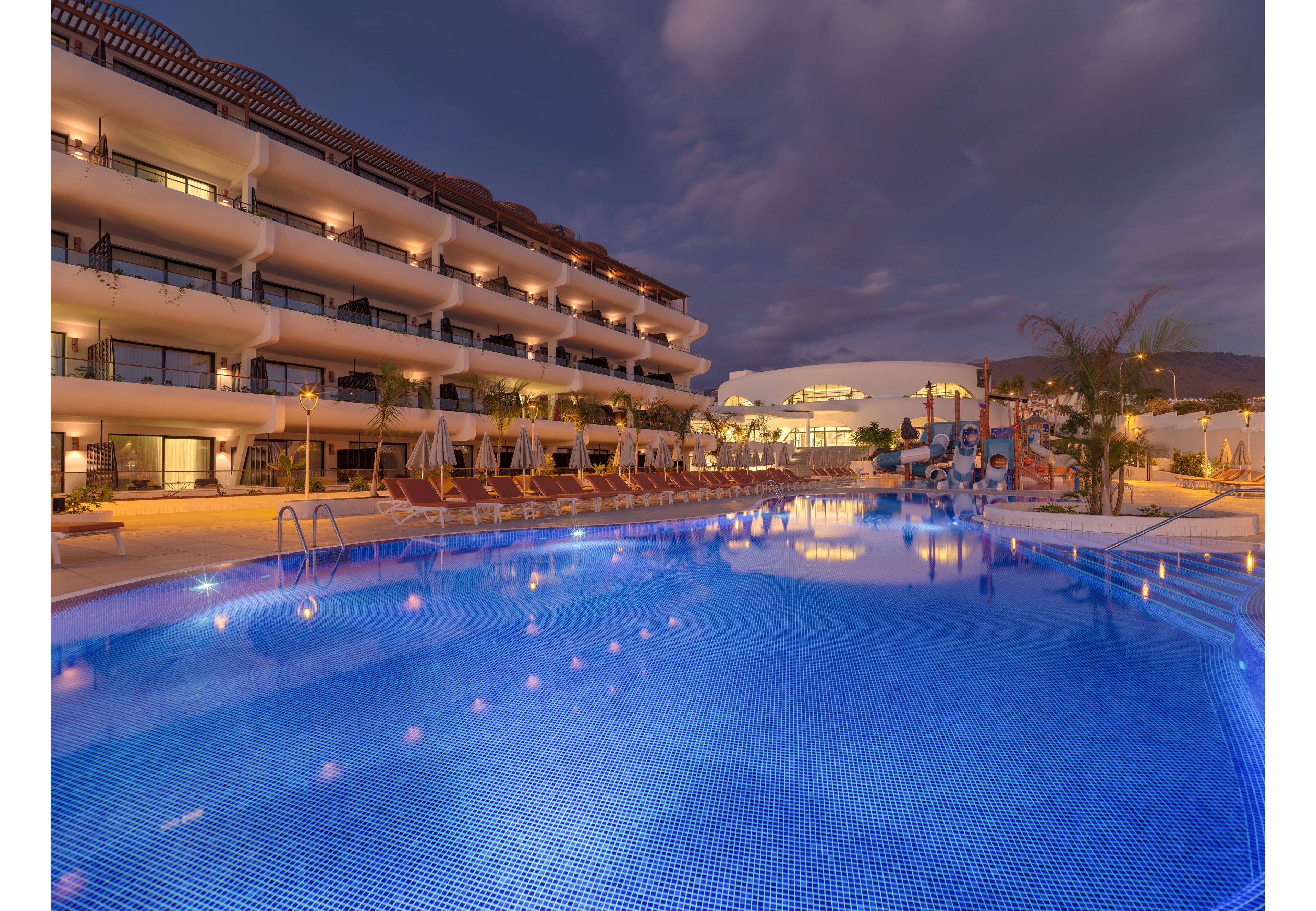 A large resort swimming pool glows with blue lighting in the early evening. The multi-story hotel building stretches along one side, with balconies overlooking the water. Rows of empty lounge chairs and umbrellas line the pool deck, suggesting a tranquil, off-peak moment. Warm architectural lighting contrasts with the cool blue pool, creating a relaxed vacation atmosphere under a cloudy sky.
