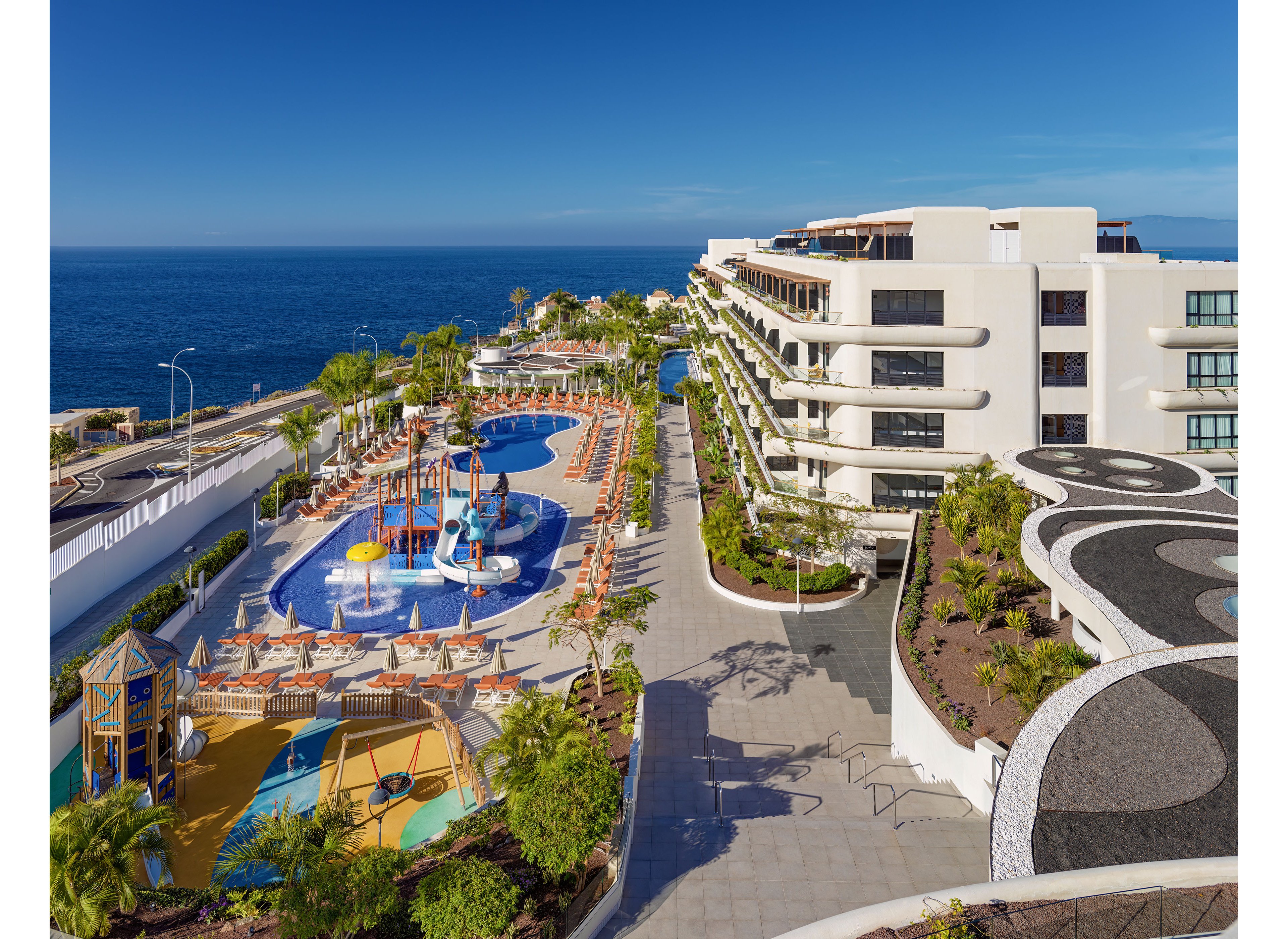 A modern seaside resort overlooks a calm blue ocean under a clear sky. Multiple outdoor swimming pools and a colorful water playground with slides and fountains are surrounded by sun loungers and umbrellas. Contemporary multi-story buildings line the right side, with landscaped paths and greenery throughout the property. The scene suggests a family-friendly vacation destination with warm, sunny weather and vibrant colors.