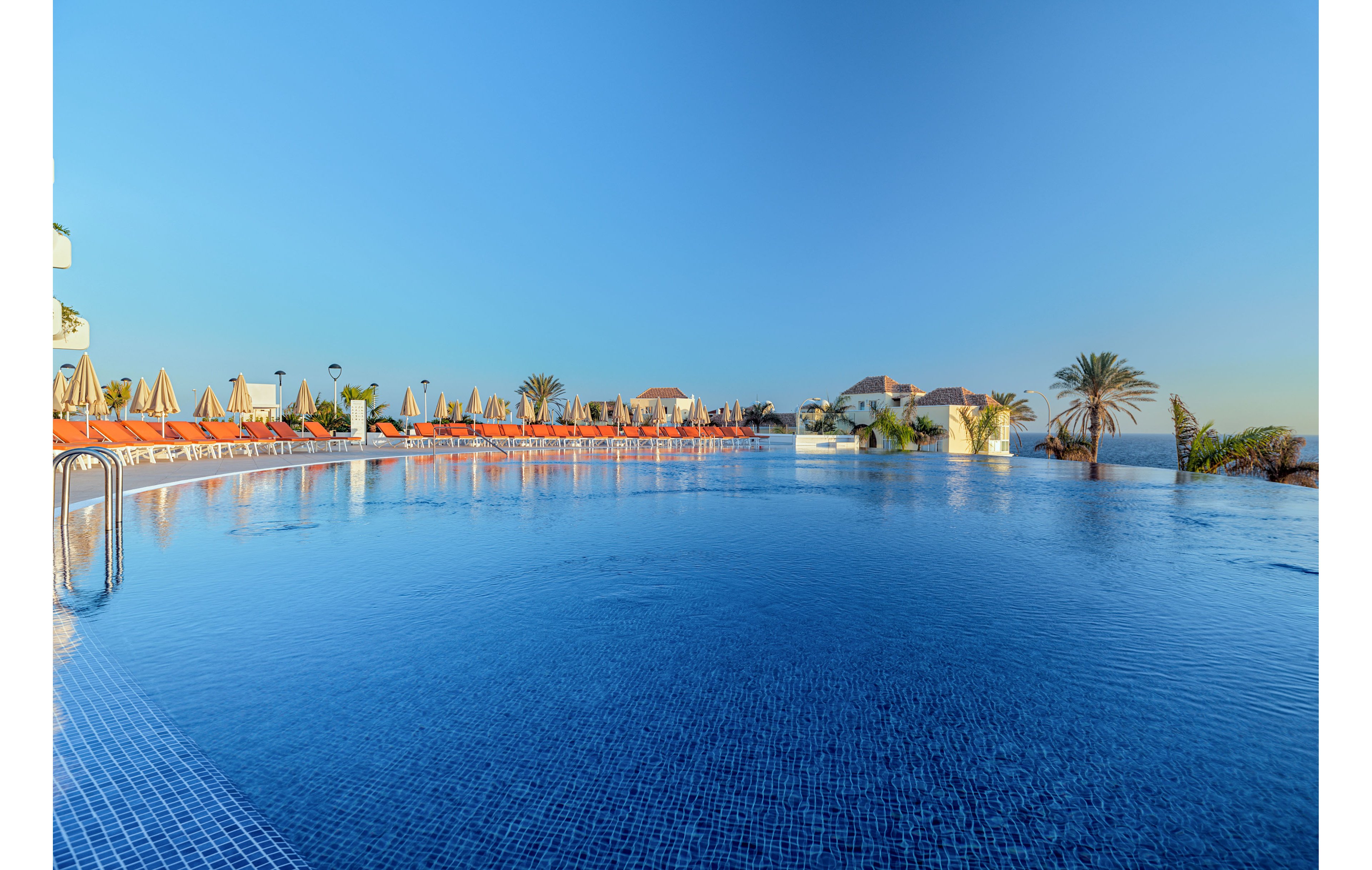 A large infinity pool stretches across the foreground, reflecting the clear blue sky. Sun loungers and umbrellas line the poolside, suggesting a relaxing resort environment. In the distance, low-rise buildings and palm trees frame the coastal setting. The calm water and warm light create a tranquil, vacation-like atmosphere.