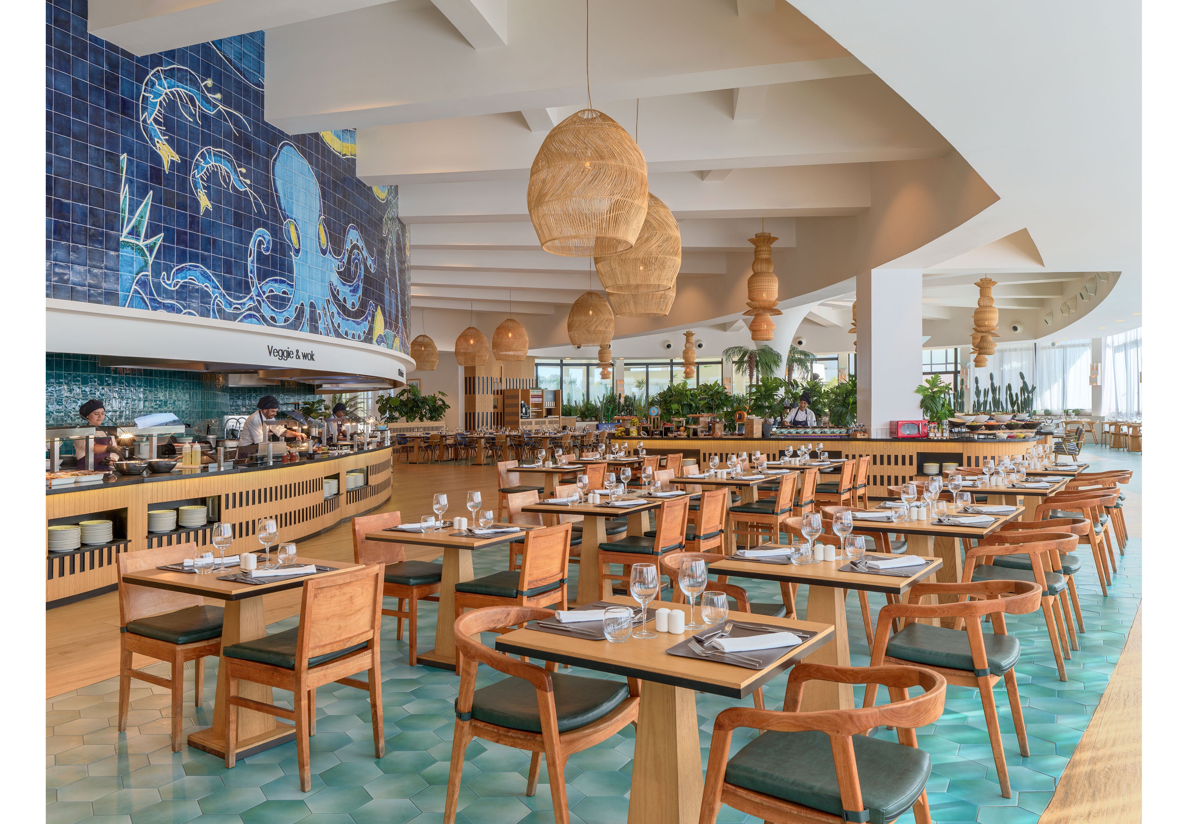 A spacious contemporary restaurant dining area features neatly arranged wooden tables and chairs with place settings of glasses, plates, and cutlery. The setting has a bright, airy atmosphere with large windows, indoor plants, and woven pendant lights hanging from the ceiling. A blue tiled wall with an illustrated octopus mural adds a playful coastal touch near the buffet counter, where the text "Veggie Land" is visible. The turquoise patterned floor and light wood accents create a relaxed, beach-inspired style.