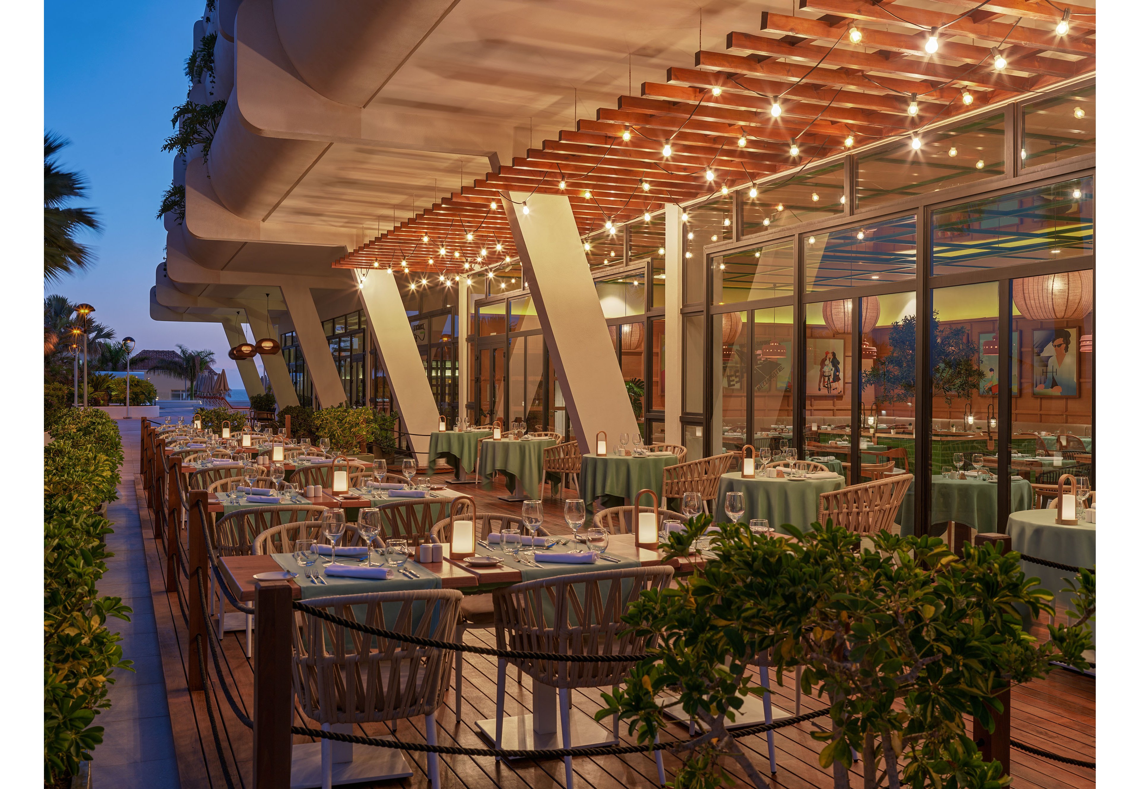 An upscale outdoor restaurant terrace is set for evening dining with neatly arranged tables and chairs. Warm string lights hang from a wooden pergola, creating a cozy ambiance along the modern building facade. Candles and table settings add a refined touch, while greenery borders the decked walkway. The sky is transitioning to dusk, enhancing the inviting atmosphere of the waterfront-style location.