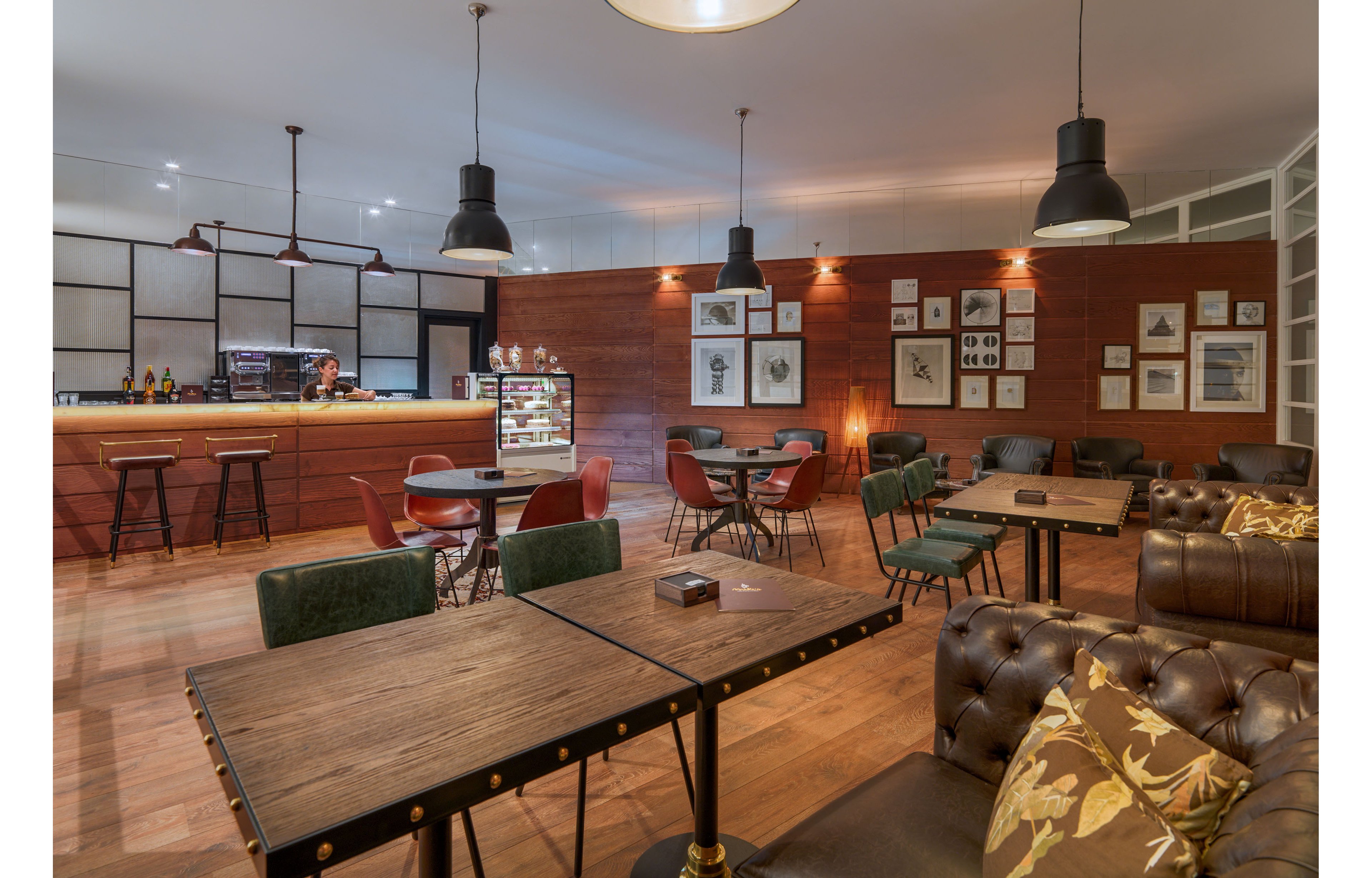 A warm, contemporary cafe interior features wooden tables and mixed seating including leather sofas and upholstered chairs. The bar area in the background displays bottles, glassware, and a lit refrigerated display case. Framed artwork decorates the wooden wall, adding visual interest and a gallery-like feel. Pendant lights hang from the ceiling, casting soft, ambient lighting over the relaxed social space.