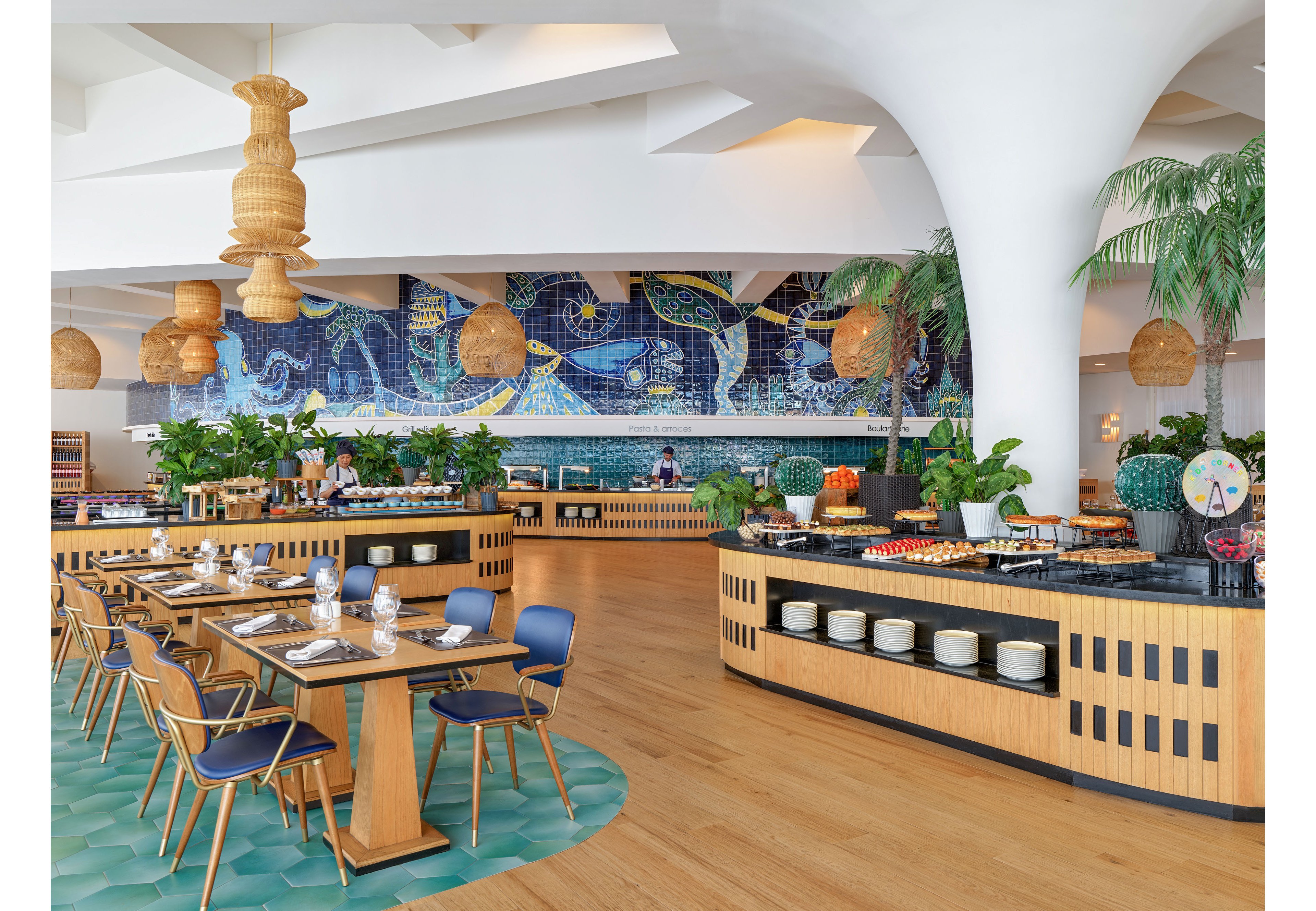 Contemporary buffet-style restaurant interior featuring wooden counters and neatly arranged dishes. A vibrant blue mosaic mural with ocean-inspired imagery decorates the back wall, adding artistic flair. Dining tables with blue chairs and set glassware occupy the foreground, while hanging wicker lamps and indoor plants create a warm, tropical ambiance. The space feels bright and inviting, with natural wood flooring and curved architectural columns enhancing the modern design.