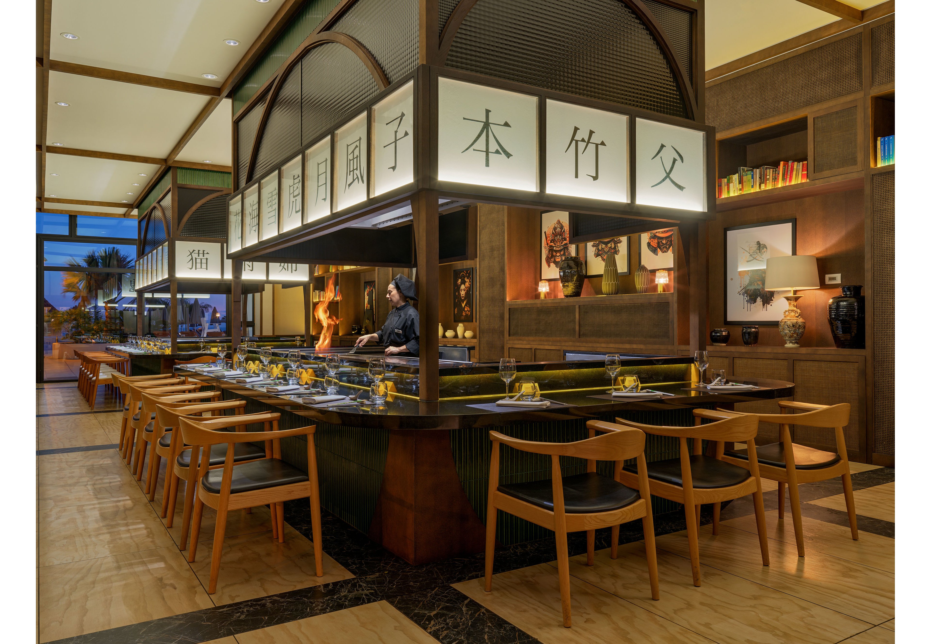 A refined Japanese-style sushi bar is shown with a chef preparing food behind a long counter. Wooden chairs surround the bar, each place setting arranged with plates, chopsticks, and glasses. Soft, warm lighting illuminates the interior, highlighting decorative artwork, shelves with books, and large vases. Overhead panels with Japanese characters add cultural detail and visual interest to the sophisticated restaurant setting.