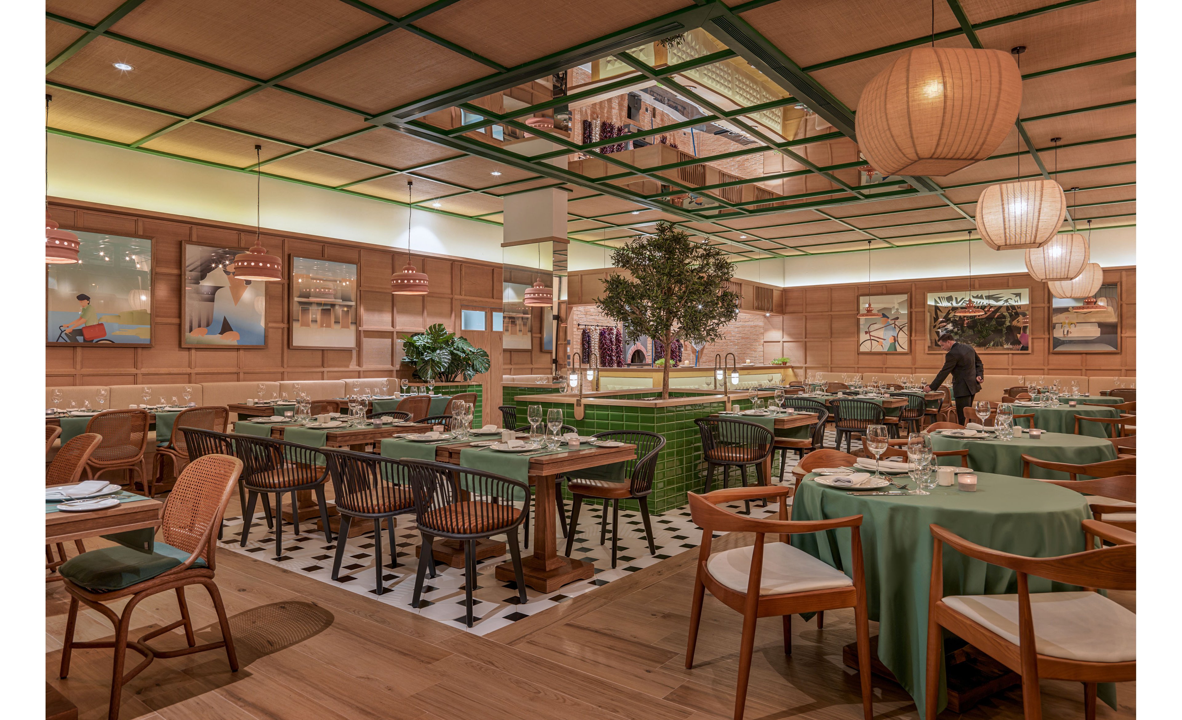 A spacious contemporary restaurant dining room features neatly arranged tables covered with green tablecloths and set with glassware and plates. Warm wooden chairs and black accent chairs surround the tables on a mix of wood and black-and-white tiled flooring. Soft pendant lighting, wall art, and a central indoor tree create a cozy, upscale atmosphere. A server walks through the space, adding a sense of activity to the elegant setting.