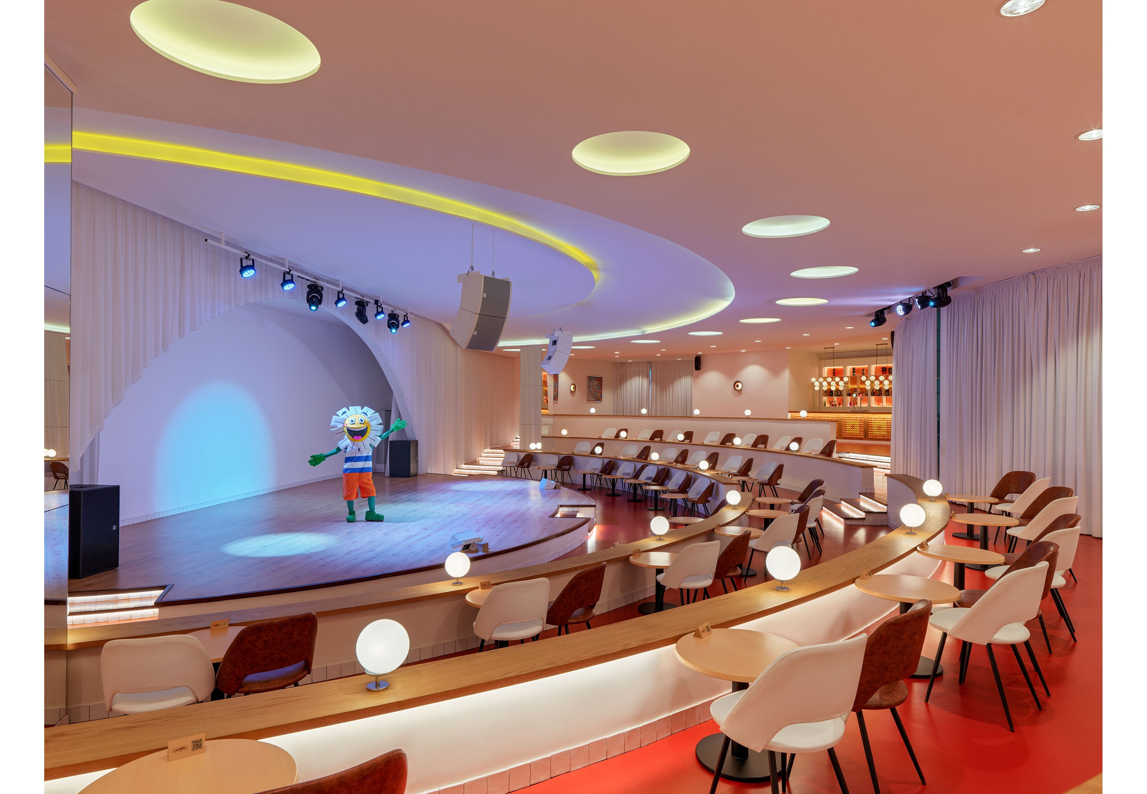 A contemporary cabaret-style theater features curved seating facing a brightly lit stage. A colorful cartoon mascot figure stands alone under spotlights on the rounded stage backdrop. The room is decorated with white chairs, warm wood accents, and spherical table lamps along the terraces. Soft pastel lighting and circular ceiling fixtures create a playful, inviting atmosphere, with a bar area visible in the background.