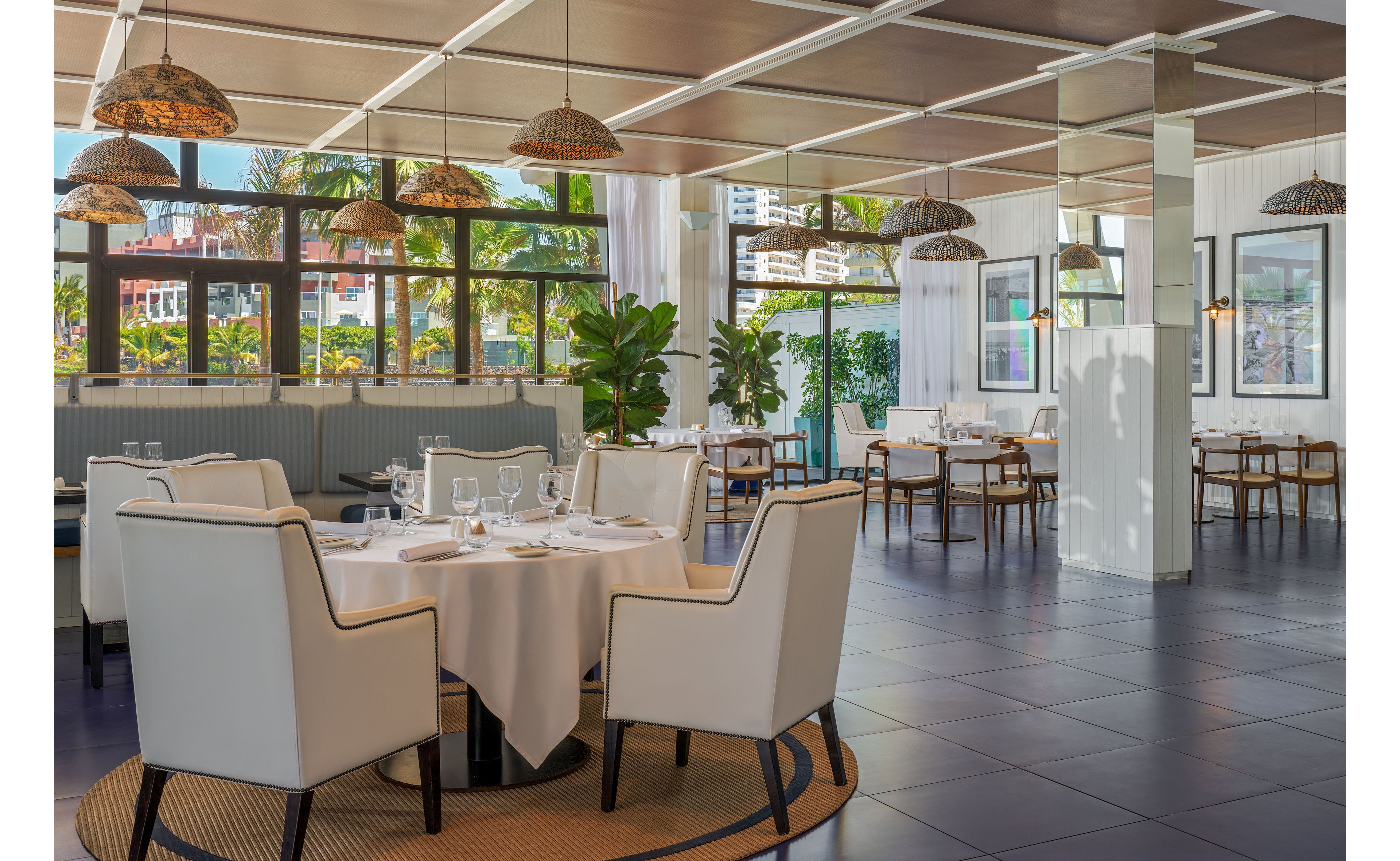 Spacious contemporary restaurant interior with neatly set round and rectangular tables. White upholstered chairs surround tables arranged on a dark tiled floor with woven rug accents. Large windows reveal palm trees and city buildings outside, bringing in abundant natural light. Green potted plants, hanging wicker lamps, and framed wall art add a relaxed, upscale coastal atmosphere.