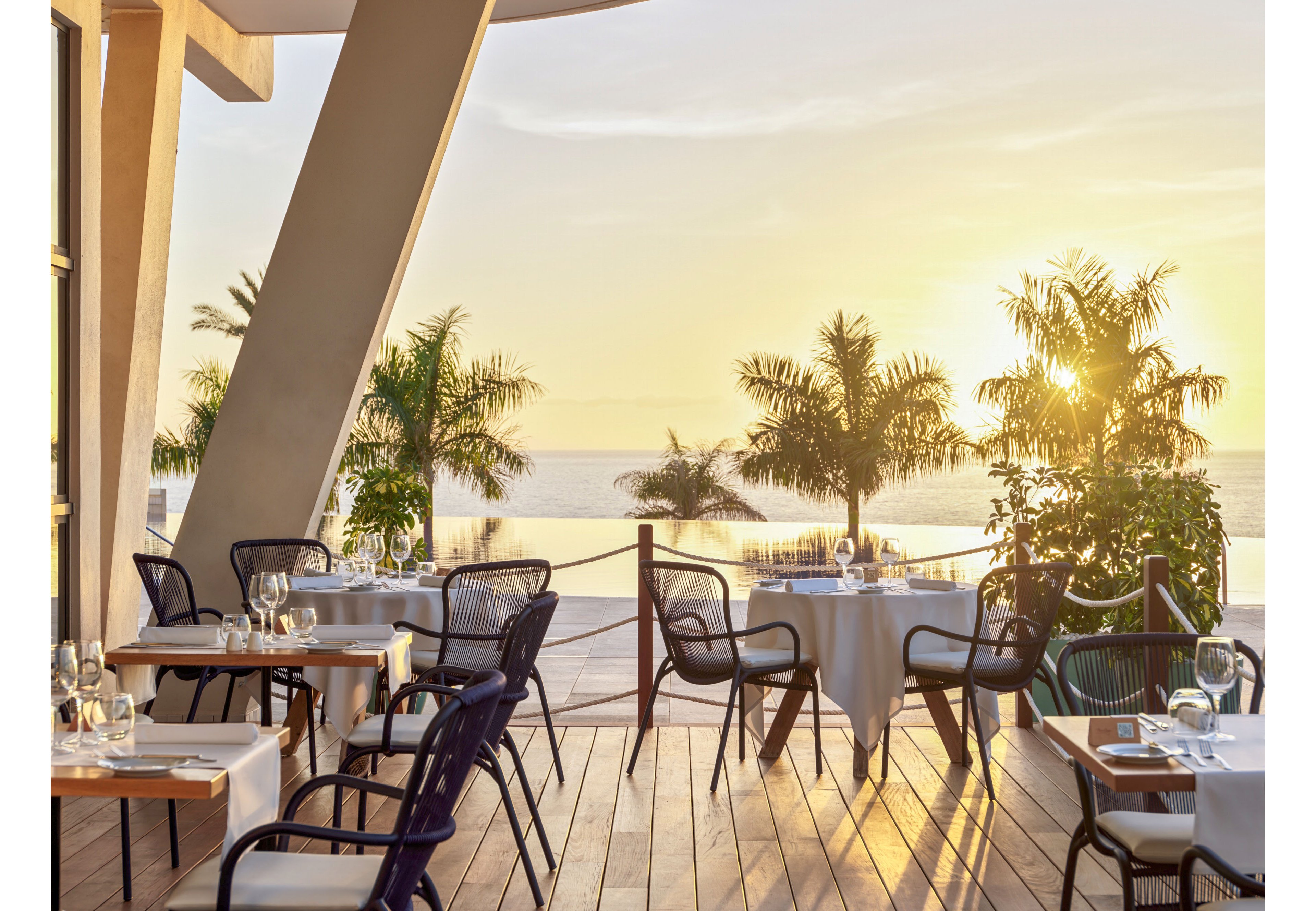 An elegant open-air restaurant terrace overlooks the sea at sunset. Tables are set with white tablecloths, glassware, and cutlery, ready for guests. Palm trees and a wooden deck with rope railing frame the view of the calm water and glowing sky. Warm golden light creates a relaxed, romantic atmosphere in this coastal dining setting.