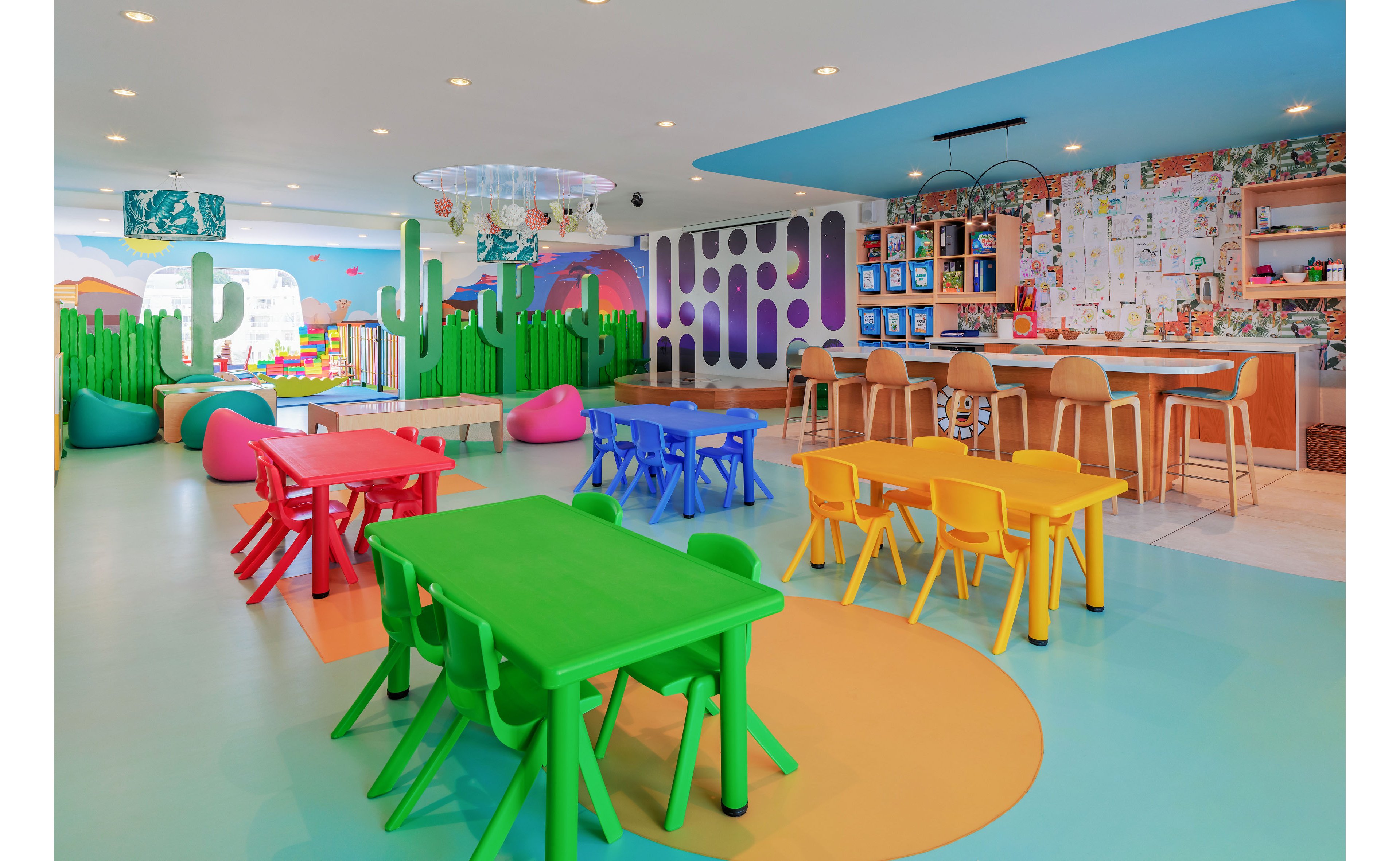Brightly colored children’s playroom features green, blue, red, and yellow plastic tables and chairs arranged on a pastel floor. The space includes playful decor such as cactus-shaped partitions, bean bags, and wall art with abstract shapes. A bar-style counter with stools and a creative wall filled with drawings and notes appears in the background. Overall, the room has a cheerful, modern design intended for kids’ activities and learning.