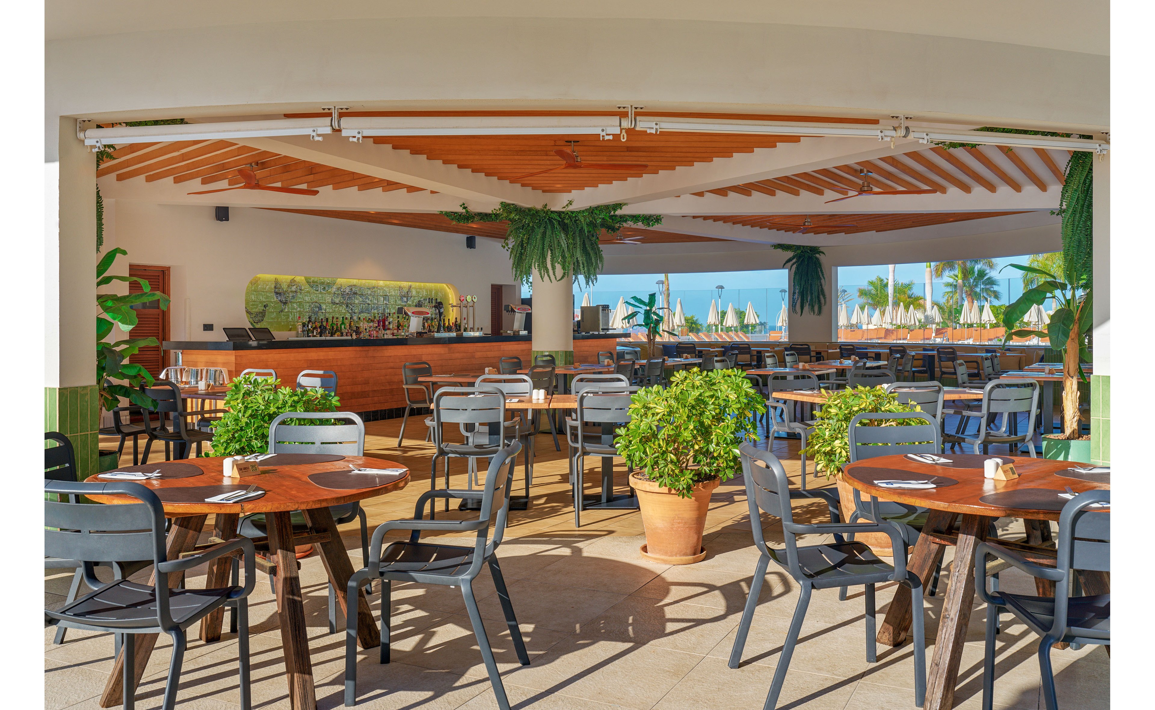 An open-air restaurant features wooden tables and metal chairs arranged on a spacious patio. Potted green plants decorate the dining area, adding a fresh touch to the bright setting. In the background, a bar counter and tropical foliage frame views of sun loungers and umbrellas by the sea. The scene suggests a relaxed coastal atmosphere ready for guests.