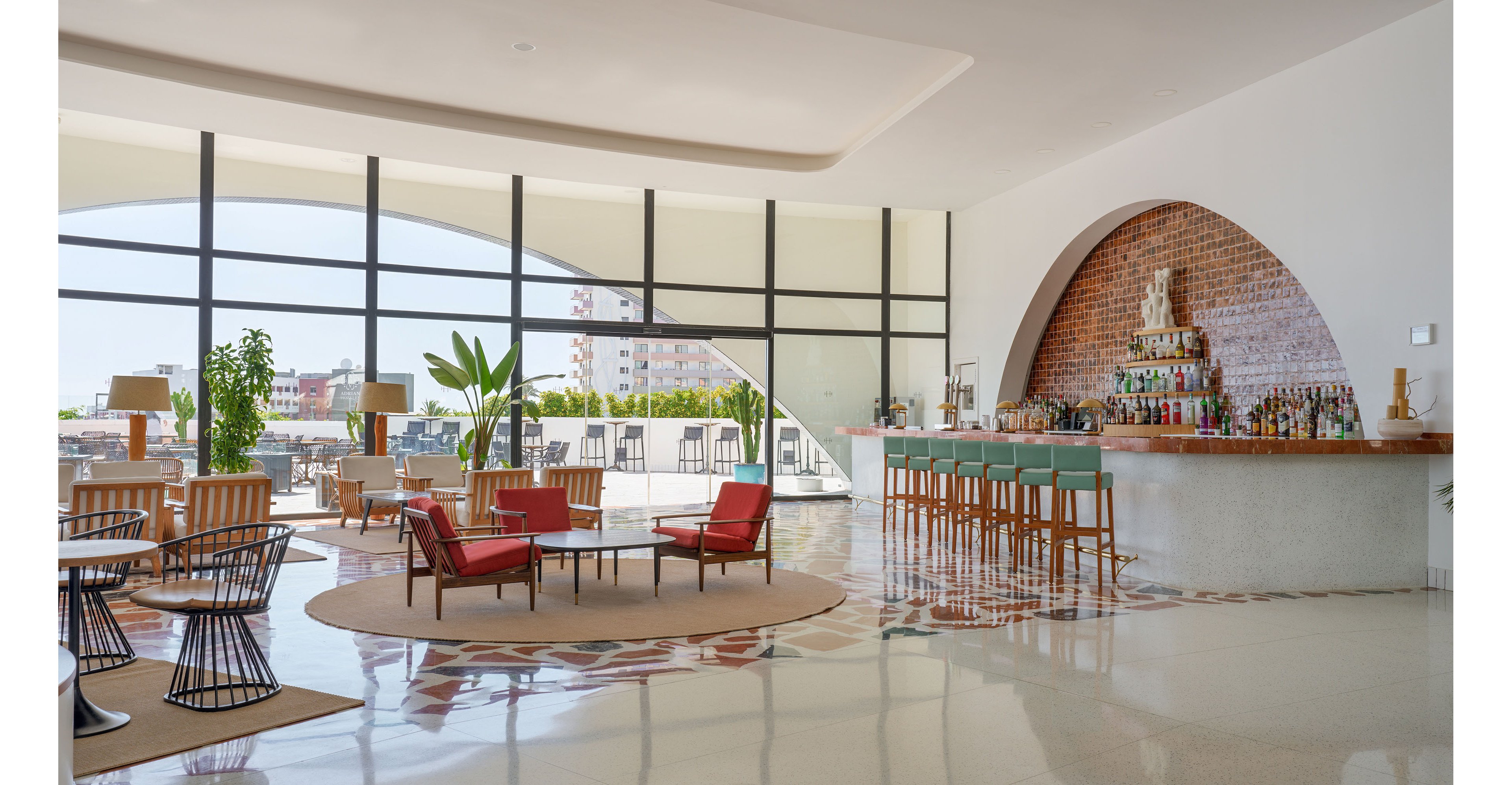 Spacious contemporary bar and lounge area with large floor-to-ceiling windows overlooking a cityscape. The scene features a curved brick-backed bar with wooden stools and colorful bottles arranged on shelves. Comfortable seating groups with red armchairs, round tables, and woven chairs are spread across a polished floor with decorative tile inlays. Potted plants and outdoor terrace furniture add a relaxed, airy atmosphere to the modern interior design.