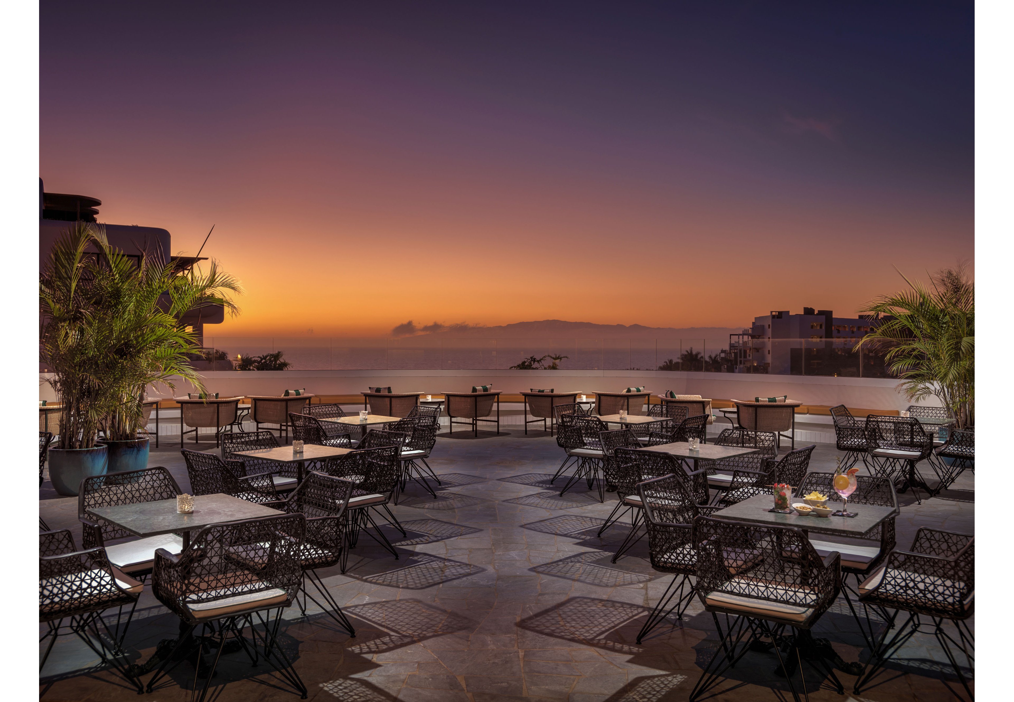 An open-air restaurant terrace overlooks the sea at sunset, with the sky glowing in warm purple and orange tones. Numerous tables and woven chairs are neatly arranged on a patterned stone floor, ready for evening diners. Potted palm plants frame the seating area, adding a tropical touch to the modern setting. In the distance, low buildings and calm water complete the tranquil coastal atmosphere.
