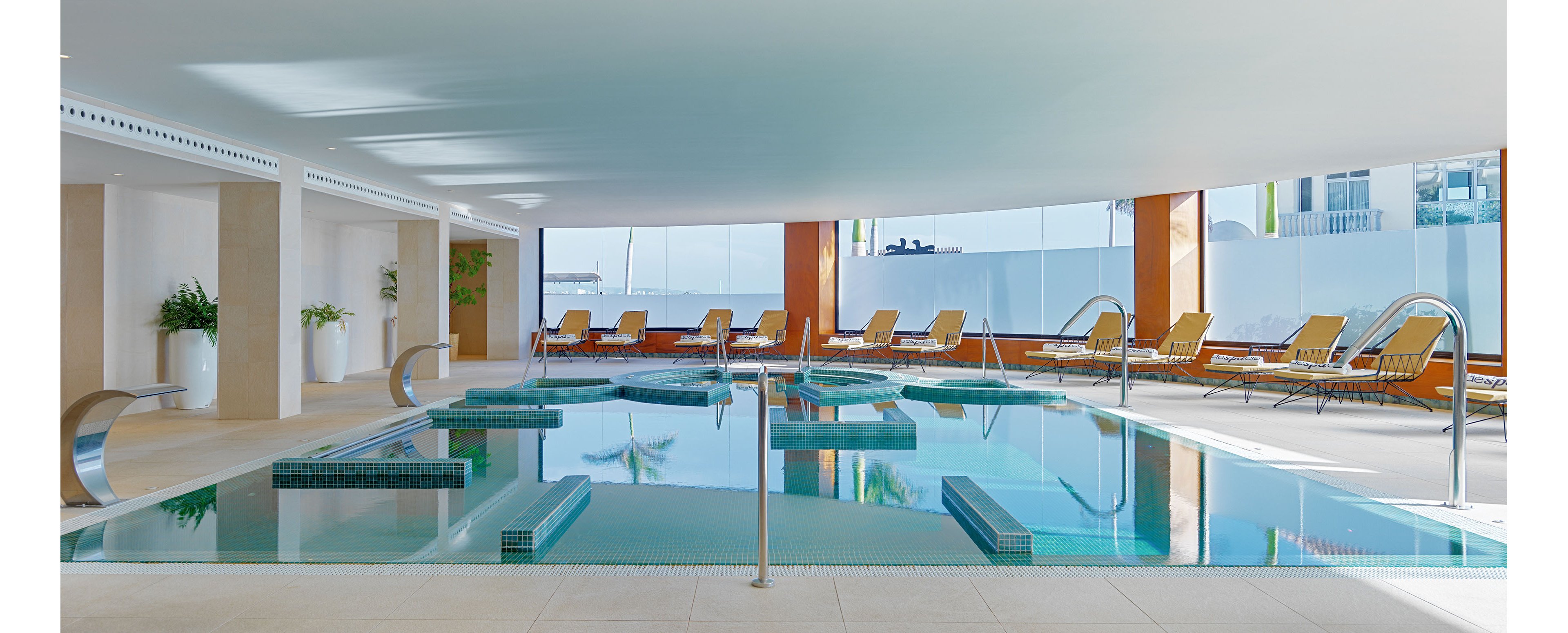 A sleek indoor spa pool features turquoise water and integrated circular seating areas. Stainless steel water features and handrails accent the contemporary design. Sun loungers line the far side of the pool, facing large windows that let in natural light and city views. The overall atmosphere is calm and luxurious, emphasizing relaxation and wellness.