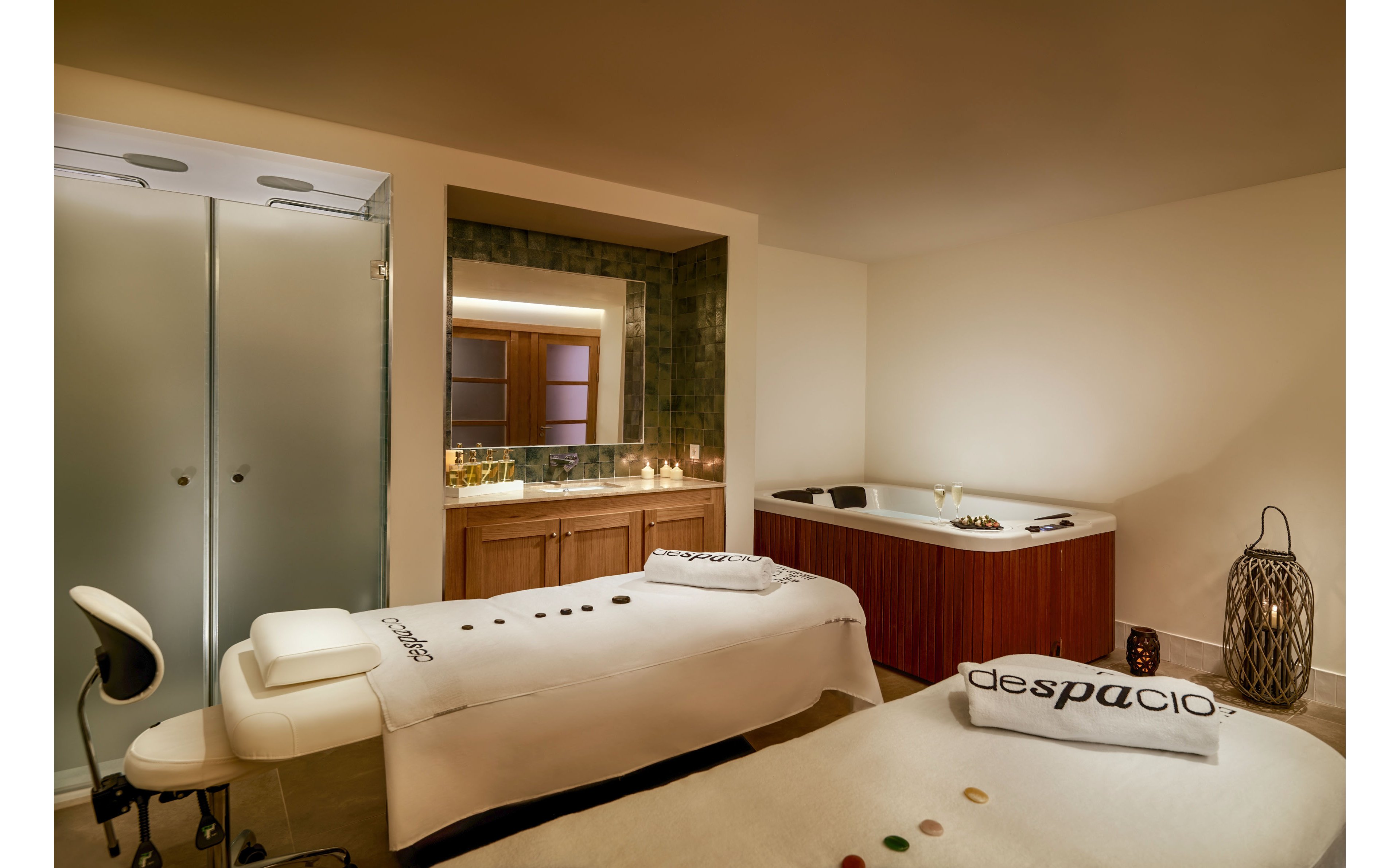 A serene spa treatment room features two massage beds neatly prepared with white linens and towels labeled "despacio." Smooth stones are arranged along the center of each bed, suggesting a hot stone or wellness therapy. In the background, a large whirlpool tub and softly lit candles create a relaxing ambiance. Frosted glass doors, warm wood cabinetry, and soft lighting enhance the tranquil, upscale atmosphere.