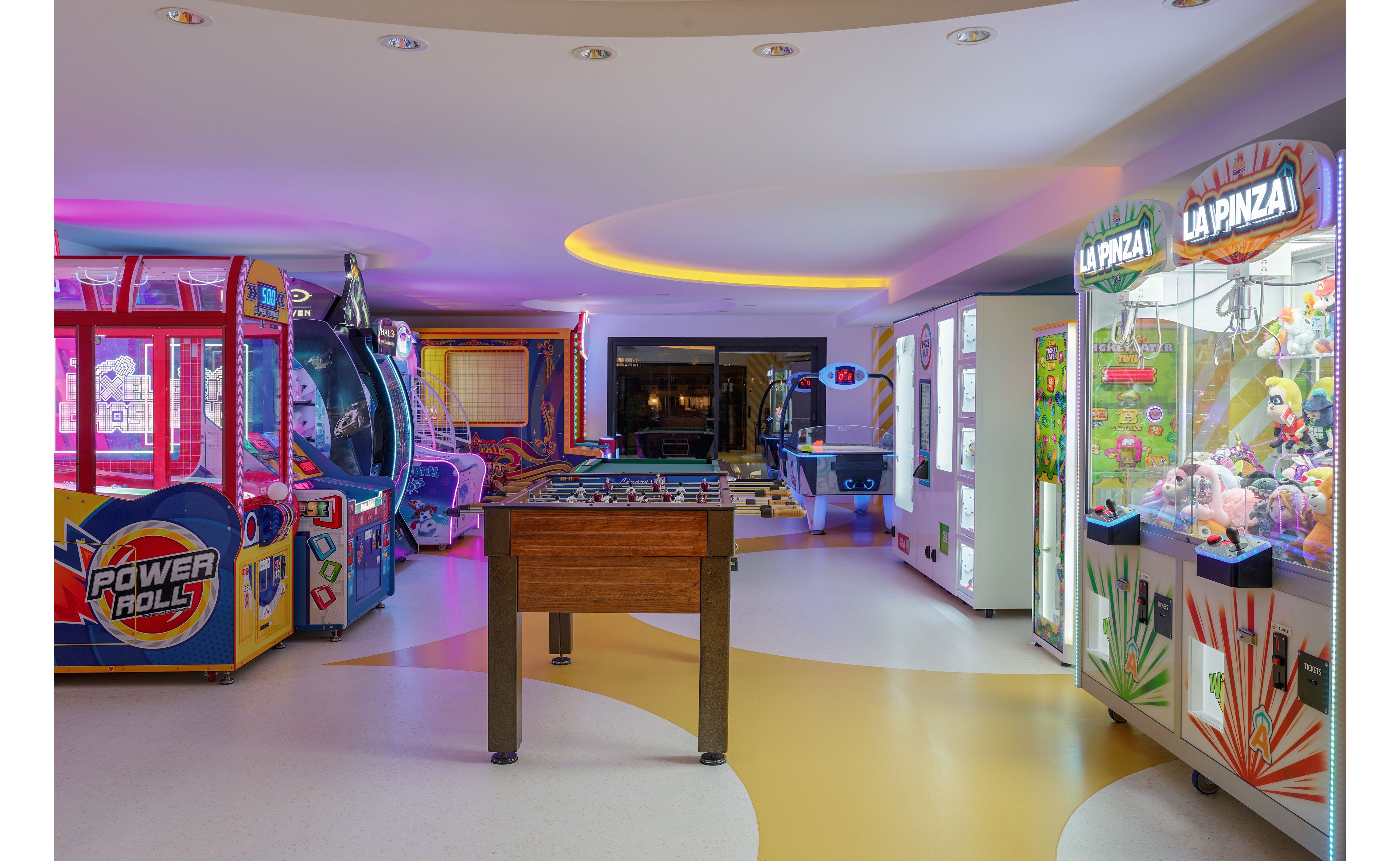 A brightly lit indoor arcade features various game machines and a foosball table in the center. Claw machines labeled "LA PINZA" and a large game cabinet marked "POWER ROLLS" stand along the sides. The space is illuminated with colorful neon and ambient ceiling lights, creating a playful atmosphere. The floor has curved patterns that guide the eye toward more games in the background.
