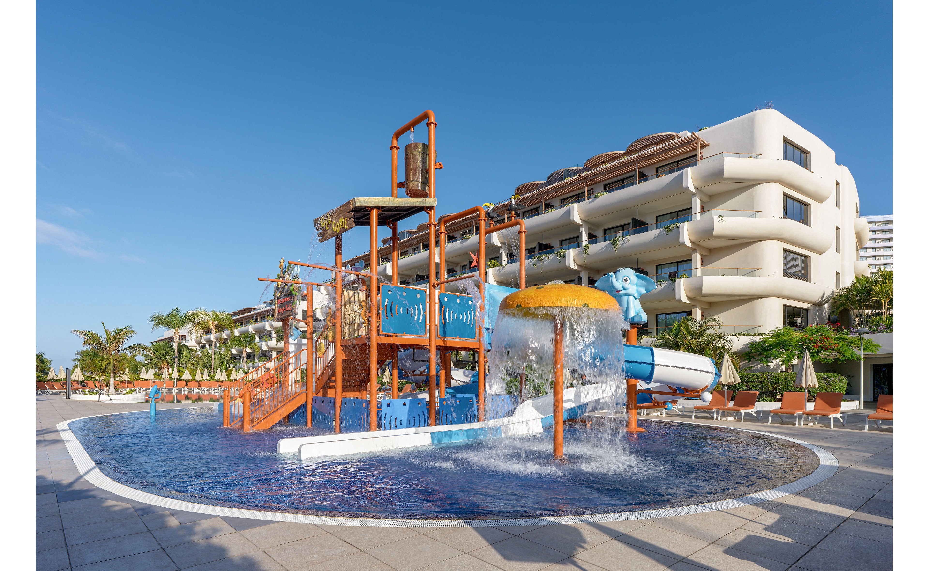 A colorful children’s water playground stands in a shallow resort pool. The structure features slides, climbing frames, and a large tipping bucket spilling water into the pool. A modern white hotel building and sun loungers with umbrellas line the background. Palm trees and clear blue sky create a bright, tropical vacation atmosphere.