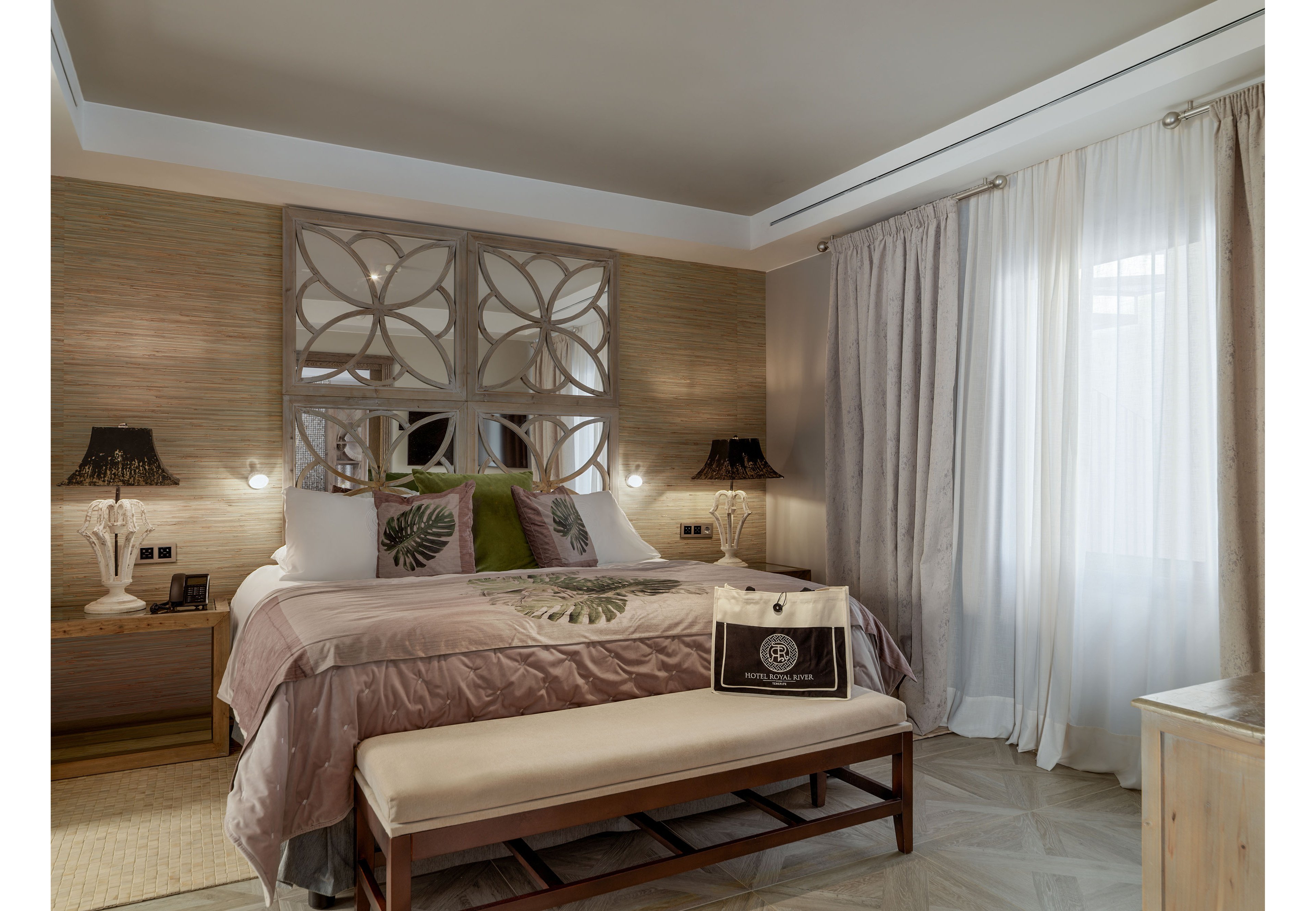 A stylish bedroom features a large neatly made bed with decorative pillows and a patterned duvet. The room is softly lit by matching table lamps on wooden nightstands, creating a warm and inviting atmosphere. A cushioned bench sits at the foot of the bed, and sheer curtains allow natural light to filter through the window. A shopping bag rests on the bench, adding a subtle touch of luxury to the contemporary interior design.