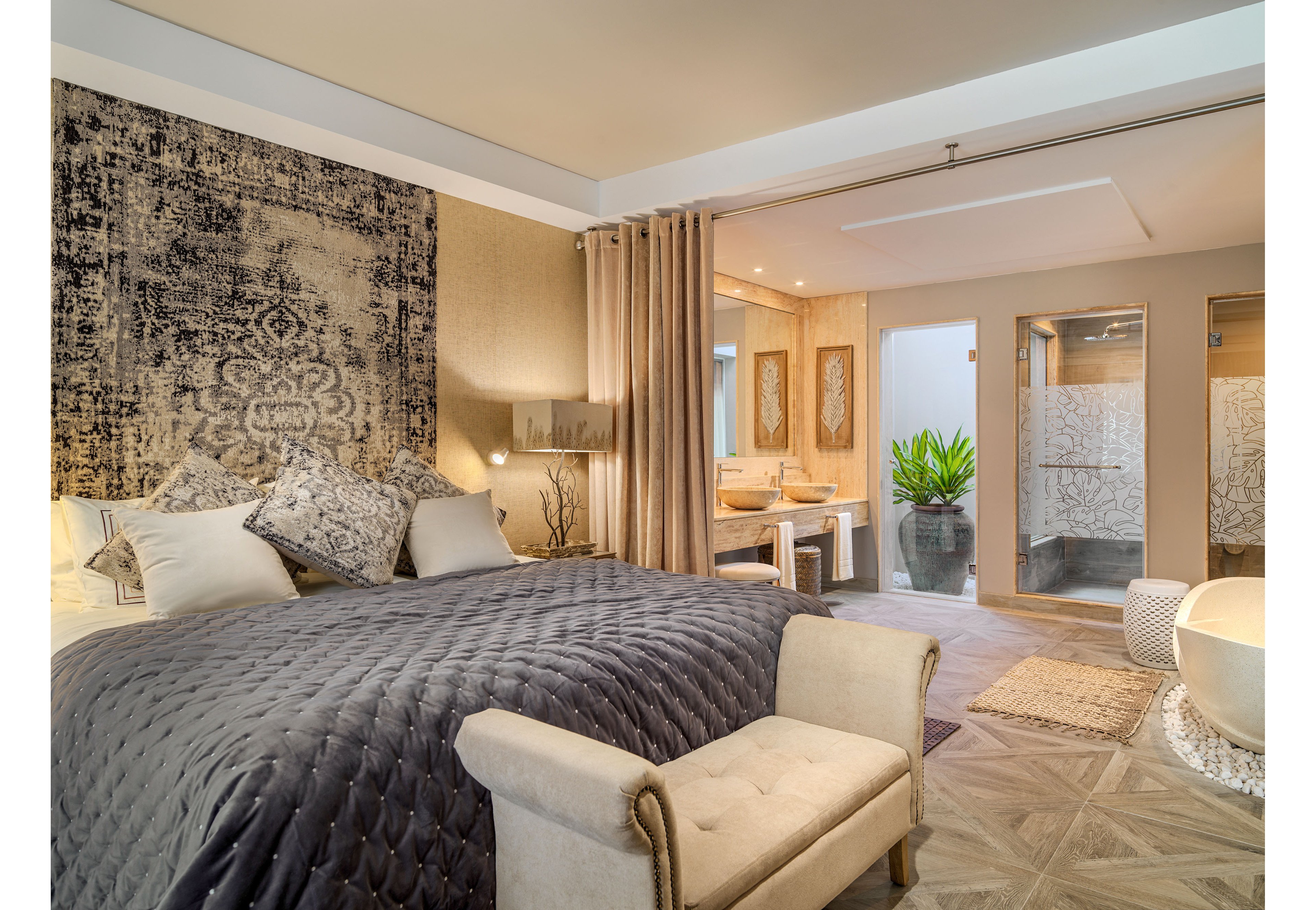 A spacious, upscale bedroom features a large bed with a quilted gray comforter and multiple decorative pillows. The room opens into an elegant bathroom area separated by curtains and glass partitions. Warm neutral tones, textured walls, and patterned rugs create a cozy yet sophisticated atmosphere. Soft lighting and a potted plant add to the relaxing, spa-like feel of the suite.